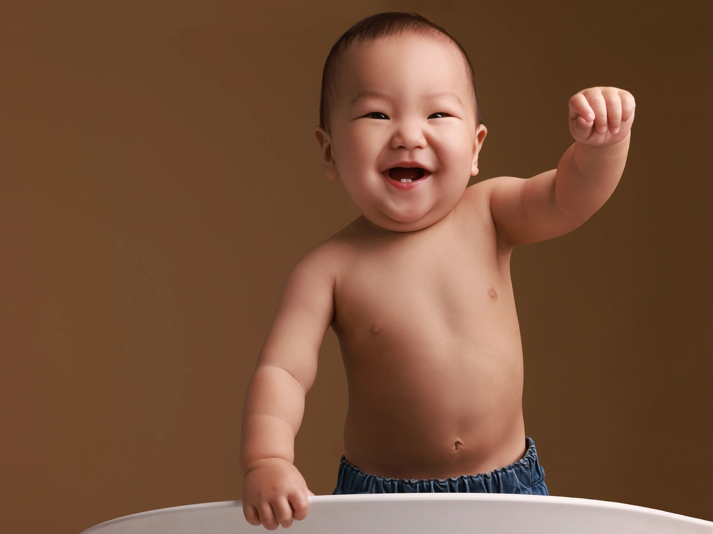 Smiling young Asian toddler with a missing tooth, raising one arm in a fist, standing shirtless behind a white surface against a brown background.