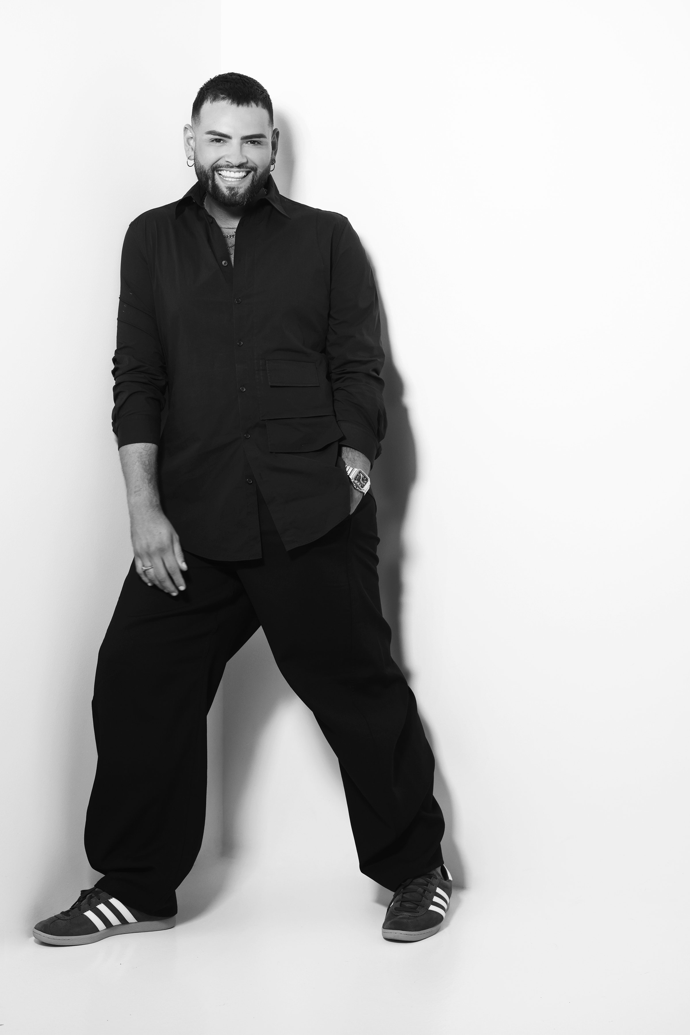 Black and white photo of a man standing against a plain white background. He has a beard, short hair, and earrings, and is smiling. He's wearing a dark long-sleeve shirt, loose dark pants, and sneakers with three stripes. His left hand is in his pock