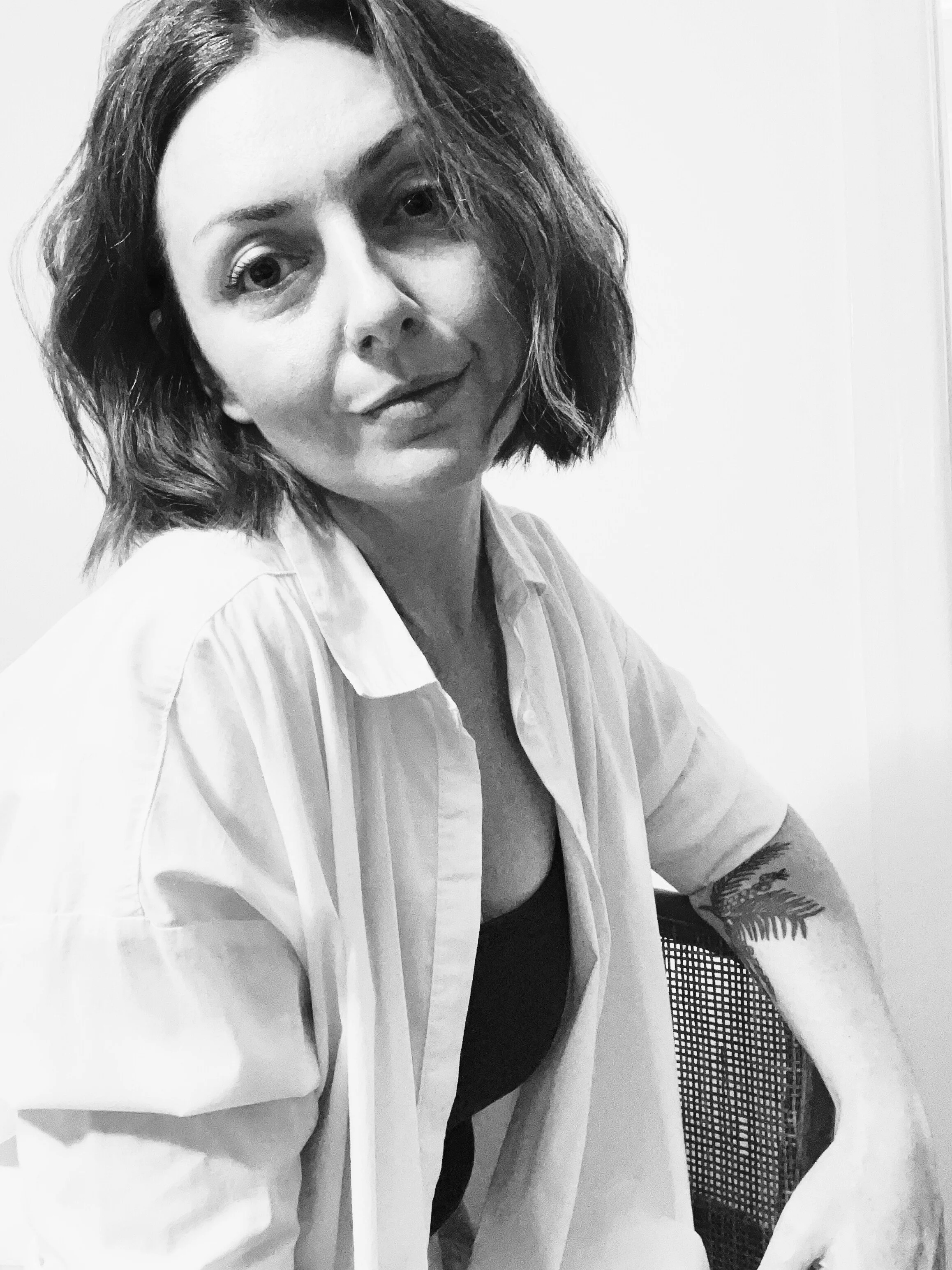 A woman with wavy, shoulder-length hair wearing a light-colored shirt and a dark top, sitting on a chair with a mesh back, looking at the camera.