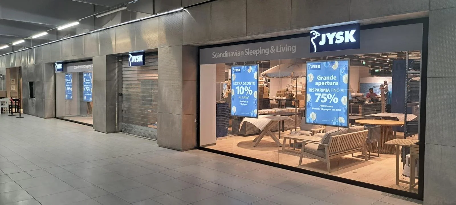 Vetrina di un negozio di arredamento JYSK con promozioni e mobili in esposizione, situato in un centro commerciale.