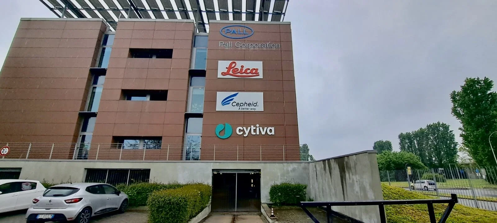 Facciata di un edificio con quattro loghi di aziende: Pall Corporation, Leica, Cepheid e Cytiva, su una parete di mattoni rossi. In primo piano ci sono alcune auto parcheggiate.