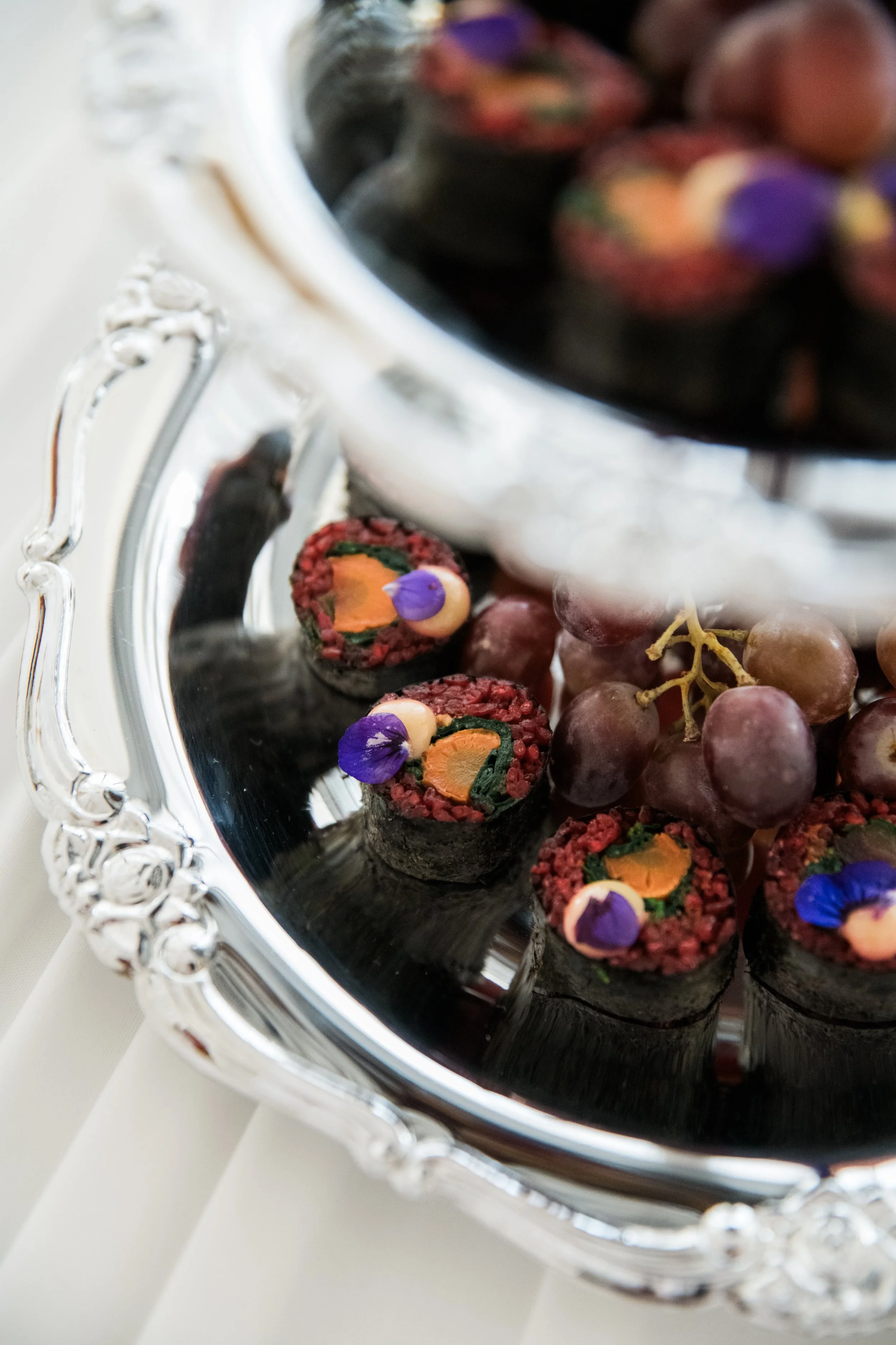 Makis au riz de betterave en feuille de nori, décorés de fleurs comestibles violettes et orangées sur plateau argenté avec raisins noirs, traiteur créatif Pays Basque, Hossegor
