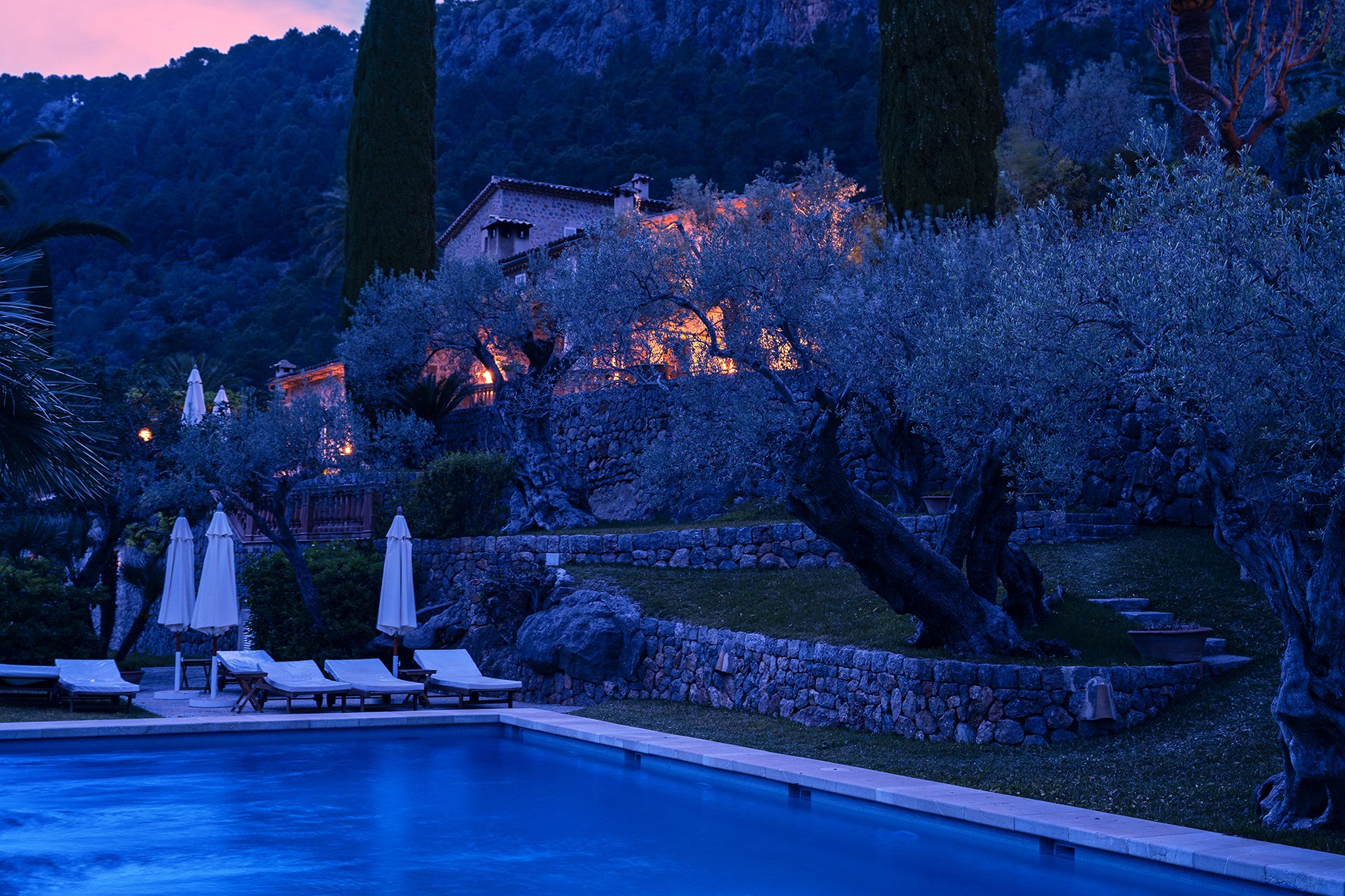 Finca Son Salas in Mallorca at blue hour seen with pool in foreground, with warm lights inside stone house, surrounded by olive trees on hillside.jpg