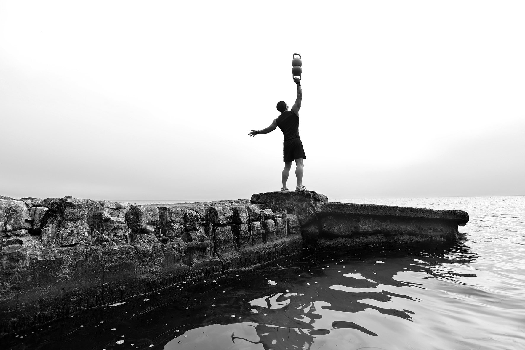Man in training outfit balancing two kettlebells on top of eachother in one arm streched overhead, on old concrete pier surrounded by water.jpg