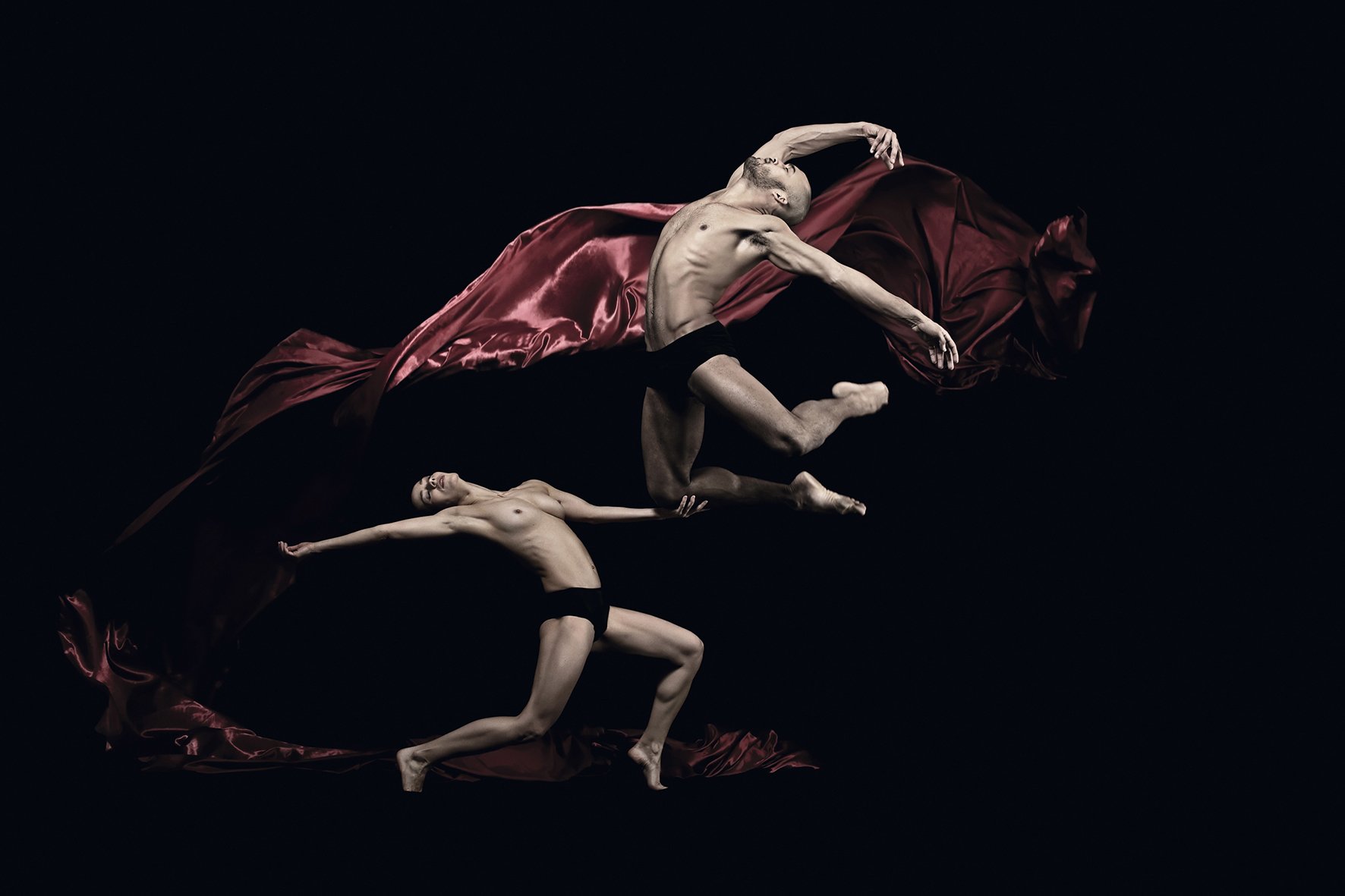 Male dancer jumping high off the ground with knees bent, while female dancer is supporting his one knee. Both semi-nude I Huge red silk fabric flying behind them.jpg