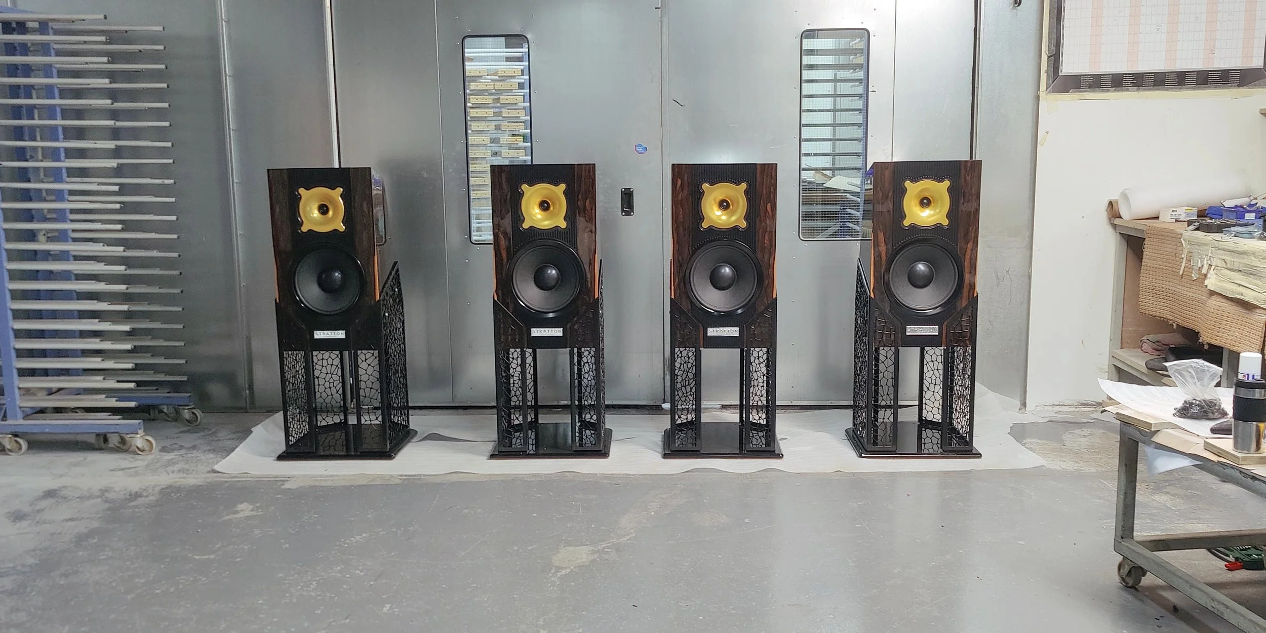 Four large black and wood Stratton Acoustics Element 12 speakers with gold waveguides are lined up on black stands in a workshop or storage room. The room has metal walls and various tools and materials on a work table to the right.
