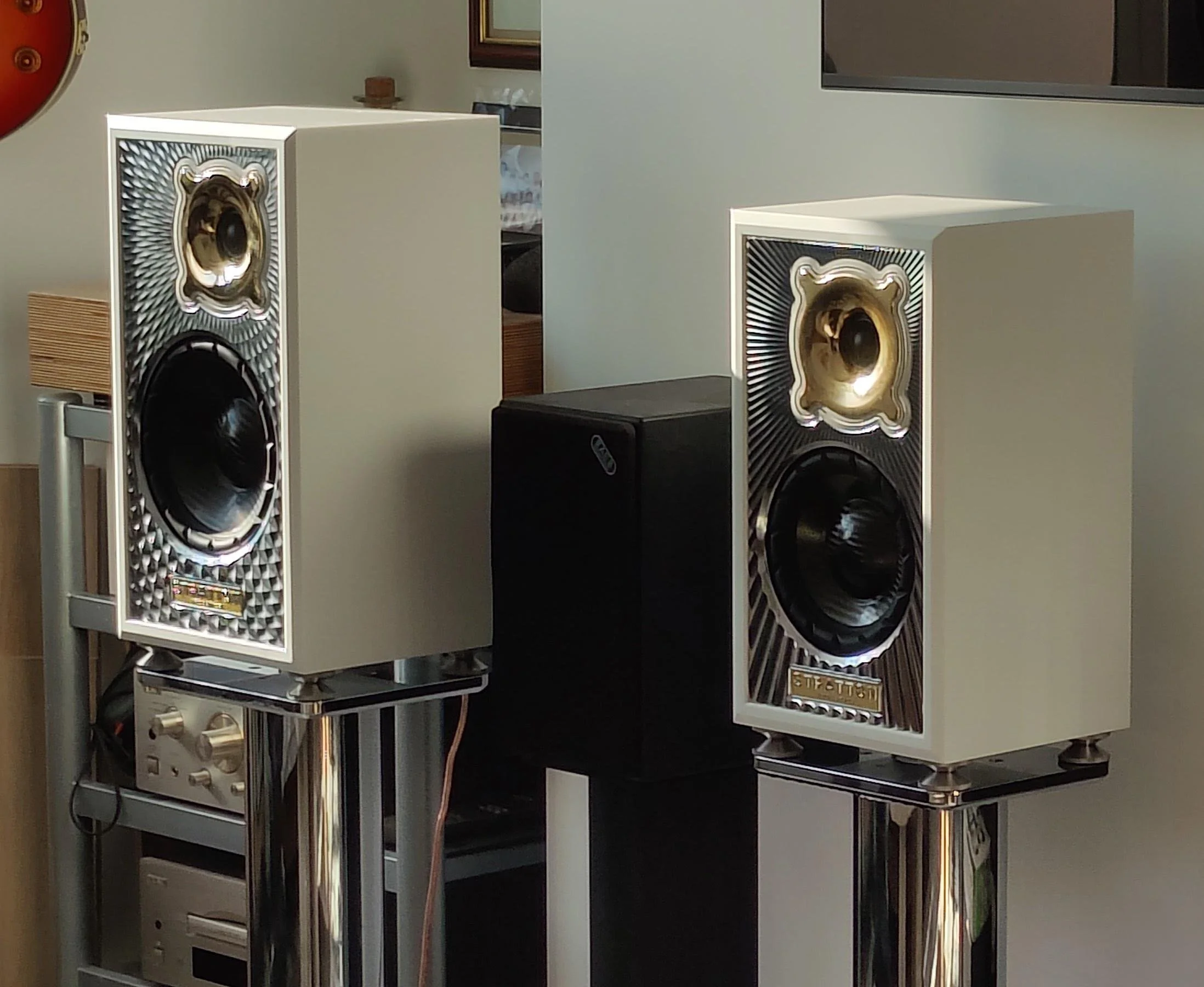 Two speakers with gold and black cones are placed on black stands, situated on a metal table with chrome legs. A smaller black speaker and a vintage stereo amplifier are also on the table. In the background, there is a wall with a mirror, a small she