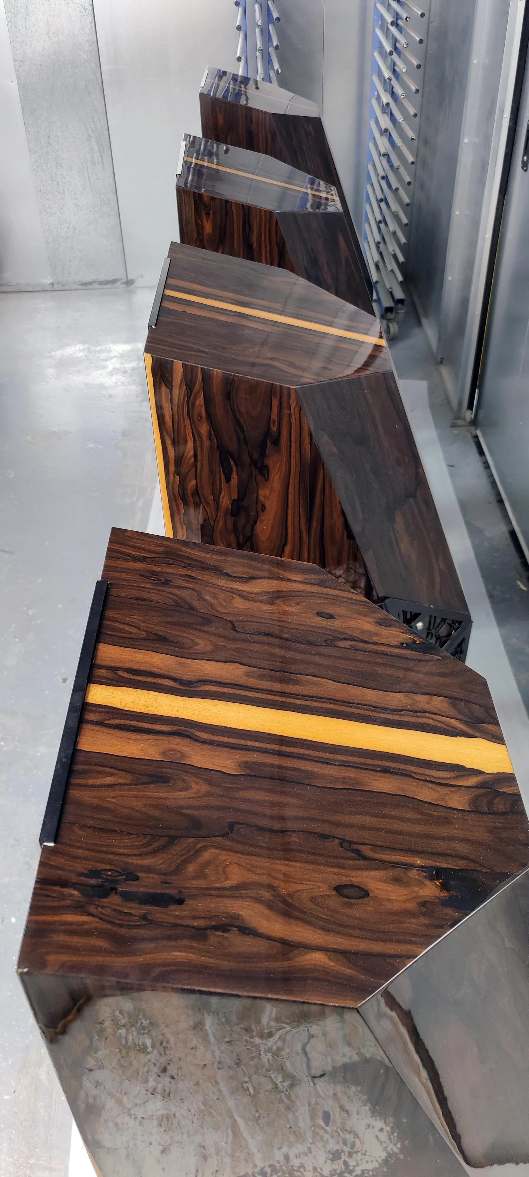 Four dark wood Stratton Acoustics Element 12 speakers with a glossy finish, showing natural wood grain, aligned in a row against a metallic wall with a metal rack or tool holder.
