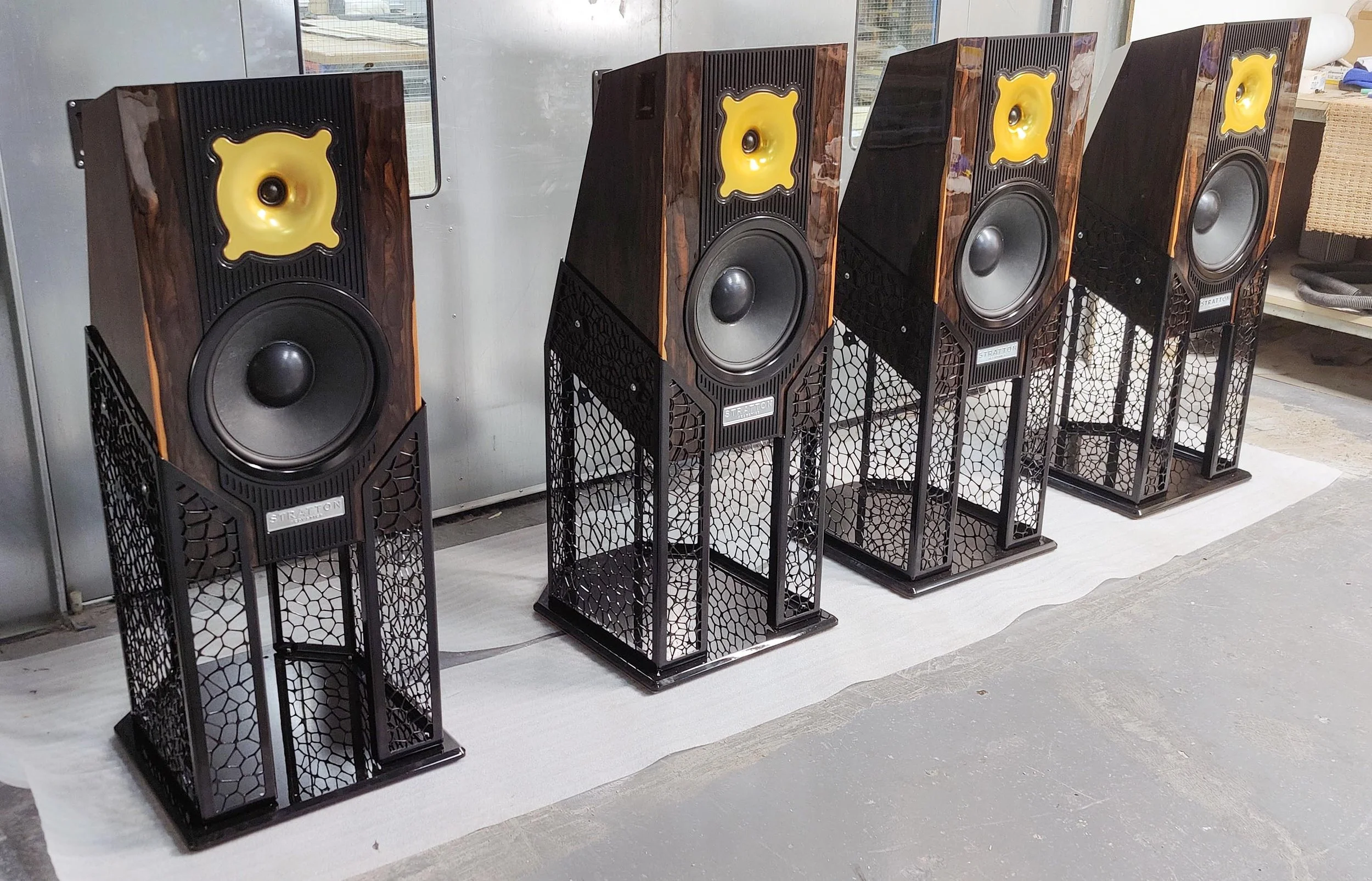 A row of large Stratton Acoustics Element 12 speakers with wooden frames and gold cones, placed on black stands with mesh sides, in a room with a concrete floor and a mirror in the background.