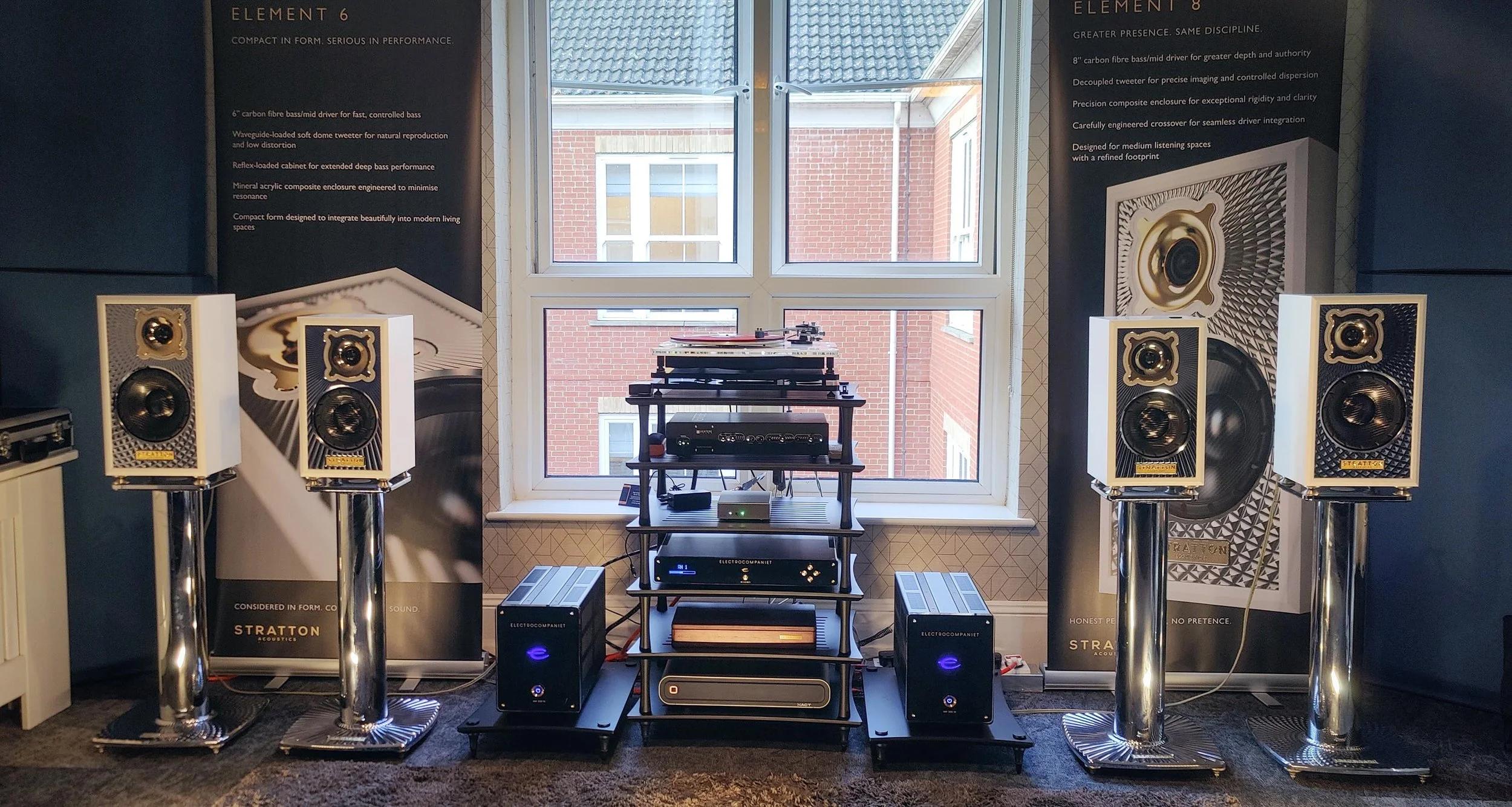 A collection of Stratton Acoustics speakers on stands, with a turntable and audio equipment set up on a black shelving unit in front of a large window with a brick building visible outside.
