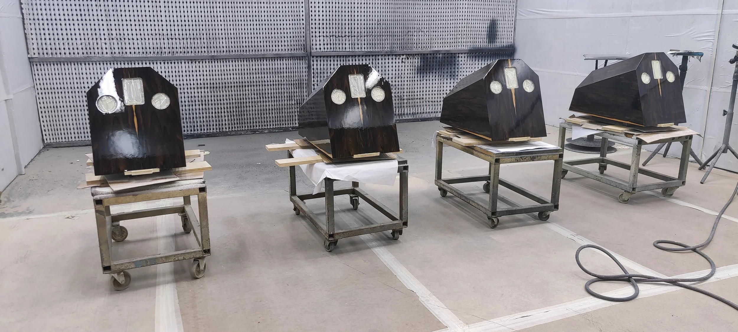 Four Stratton Acoustics Element 12 speakers are placed on metal carts in a workshop. They have dark, polished wood with light-colored circular and rectangular elements.