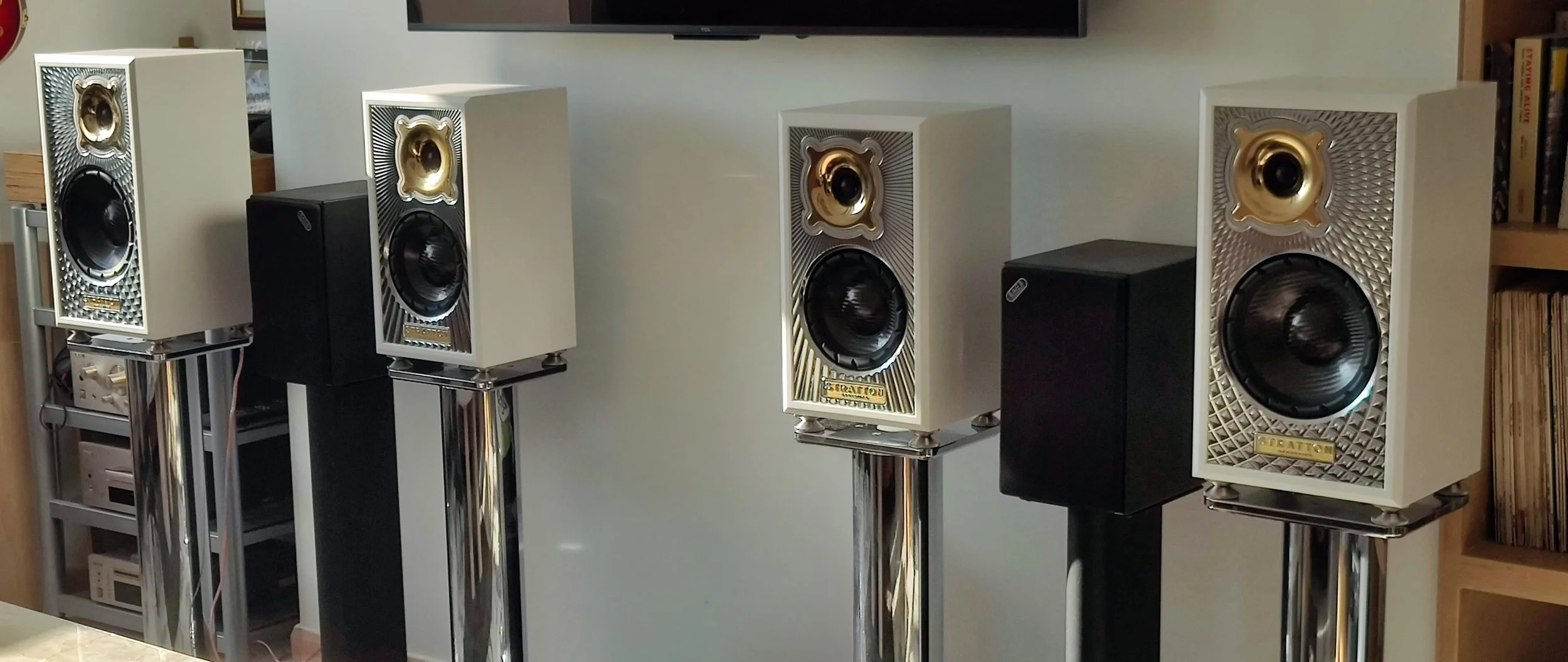 Collection of white and black loudspeakers with gold accents on tall, shiny stands in a room.