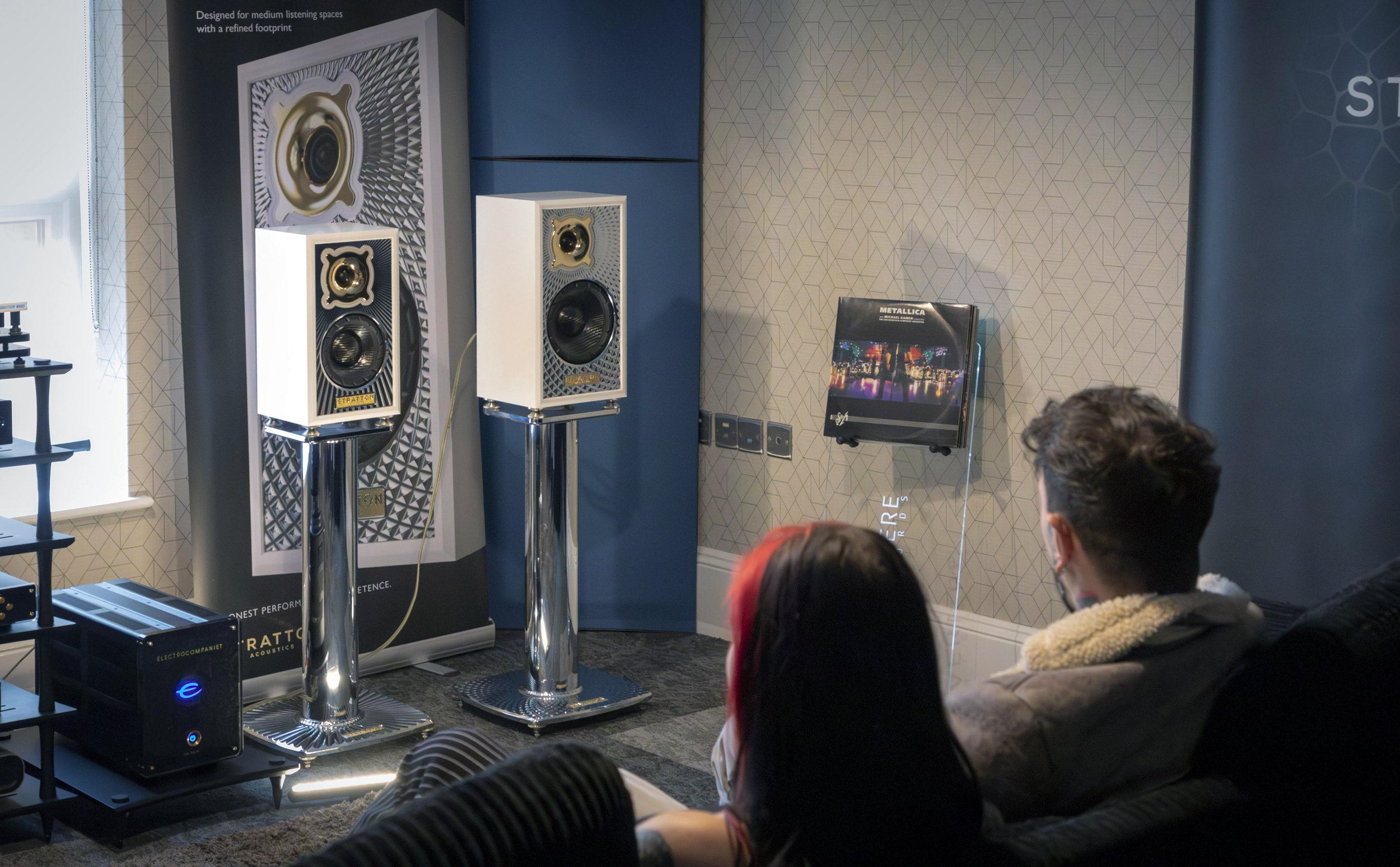 A group of people sitting in an audio equipment show, observing and listening to high-end speakers and audio gear displayed on stands and walls.