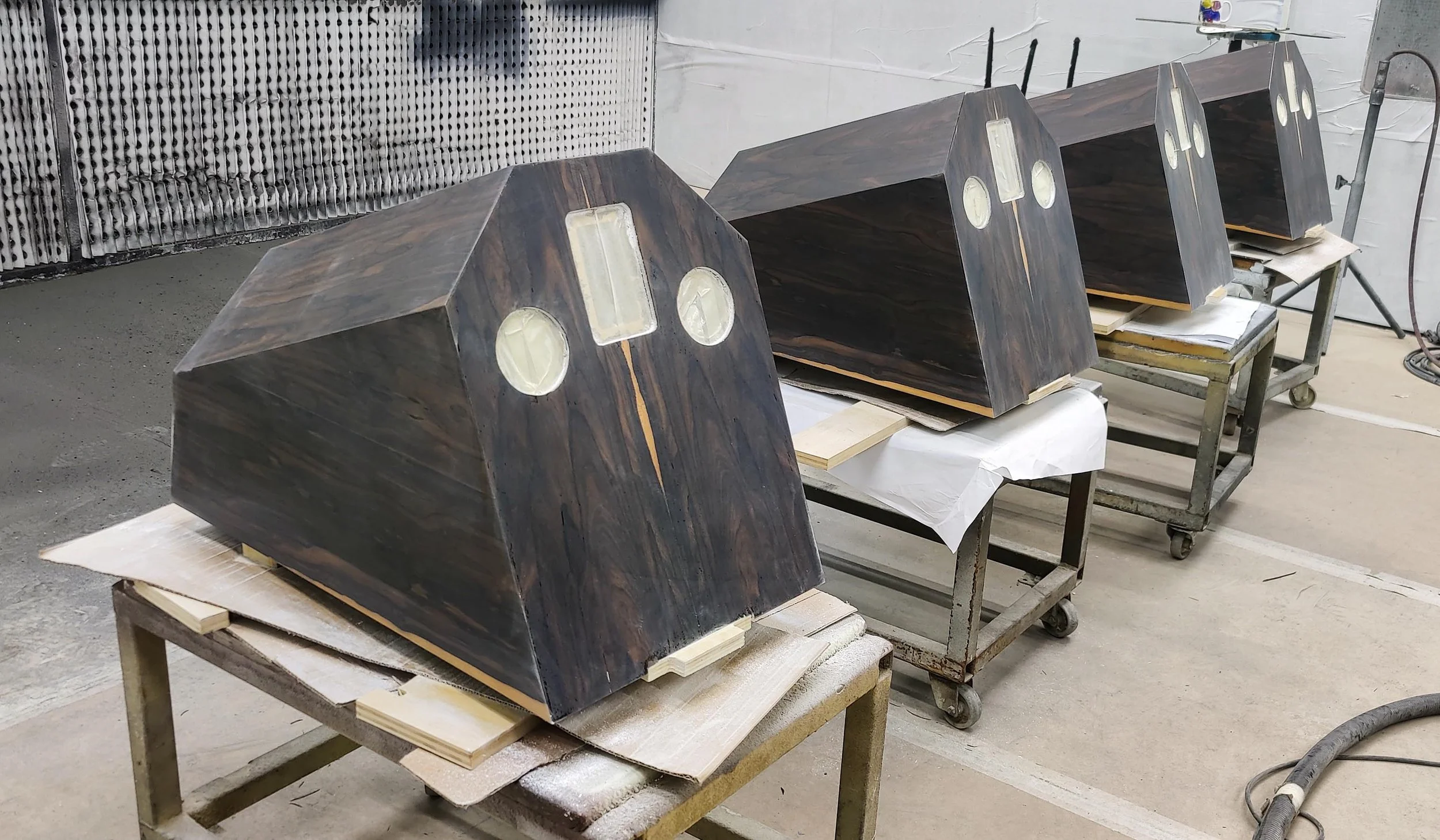 Wooden Stratton Acoustics Element 12 speaker enclosures with oval and rectangular openings in a workshop setting, placed on carts.