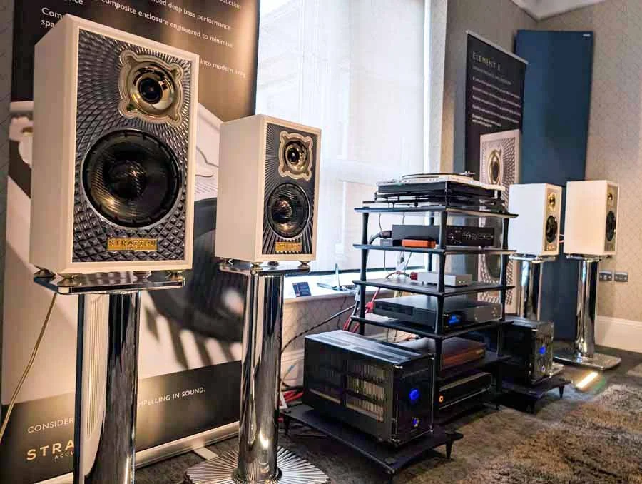 Four white Stratton Acoustics speakers mounted on chrome stands, with audio equipment and stacked components, with posters and a window in the background.