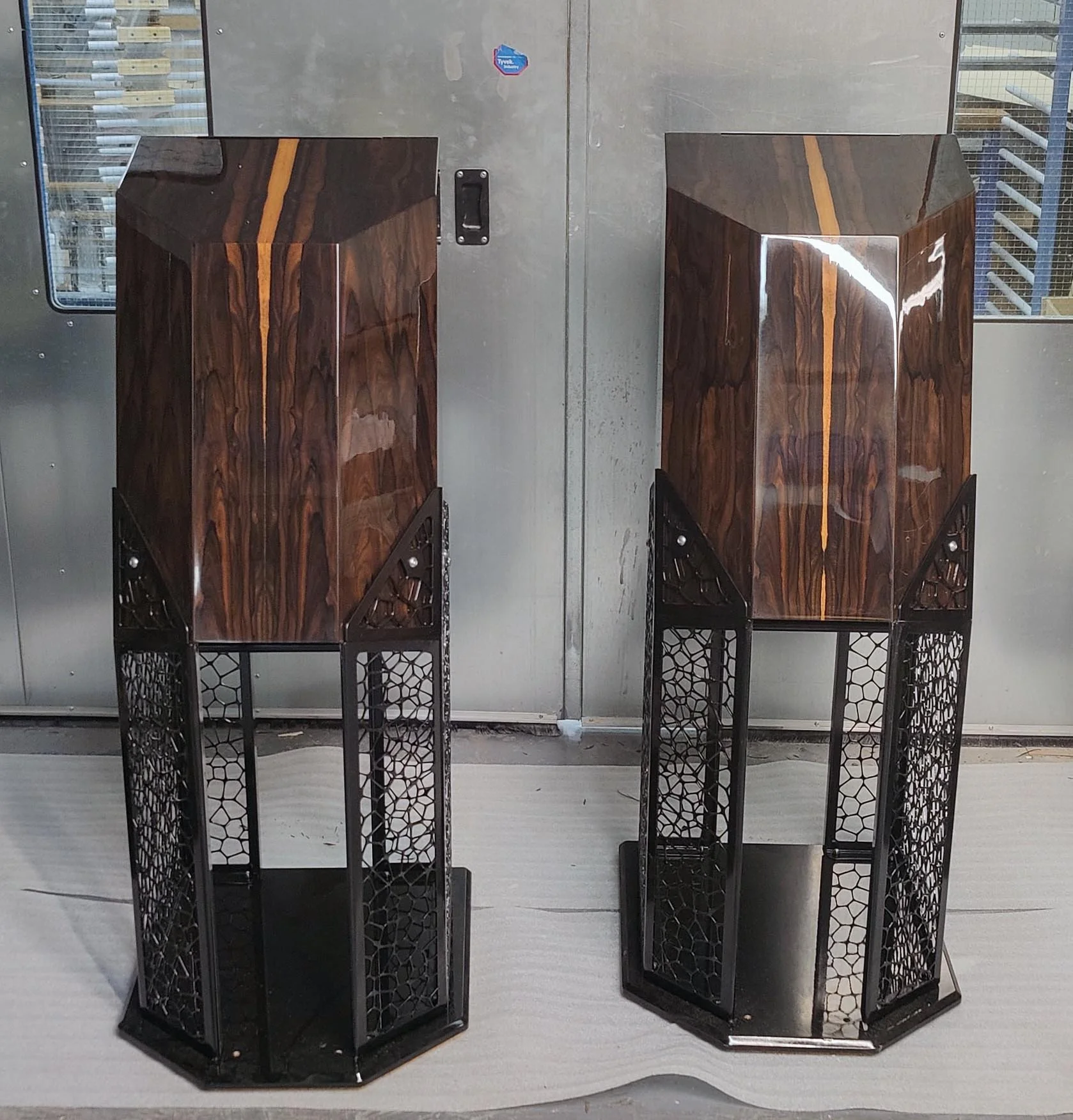 Two  with a dark wood grain finish and black metal legs are positioned in front of a metallic wall with a small sticker. To the left and right of the speakers, there are black metal stands. The speakers are symmetrical, with the same design and size.