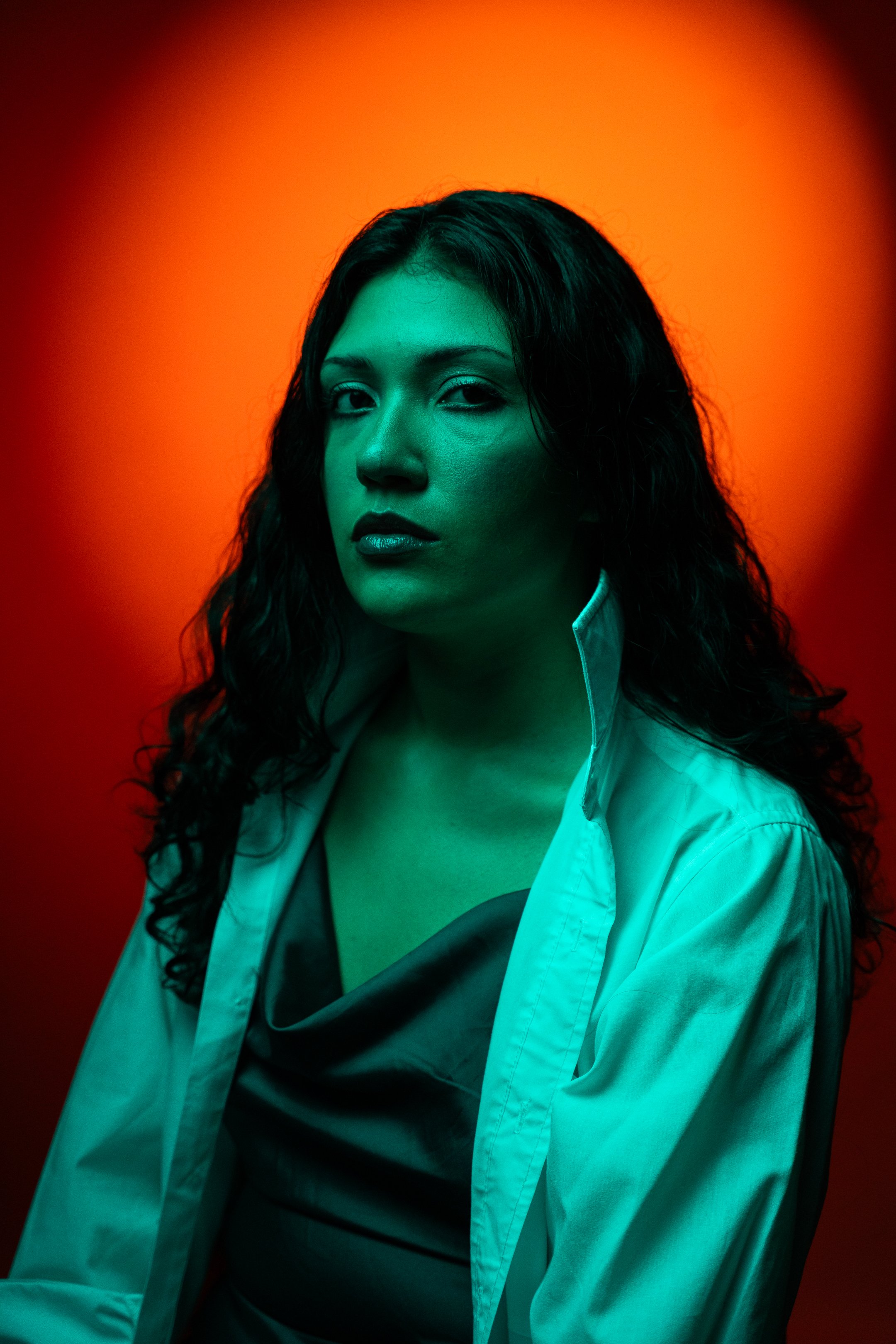 Portrait of a woman with dark curly hair wearing a white shirt and black top, illuminated with teal and orange lighting.
