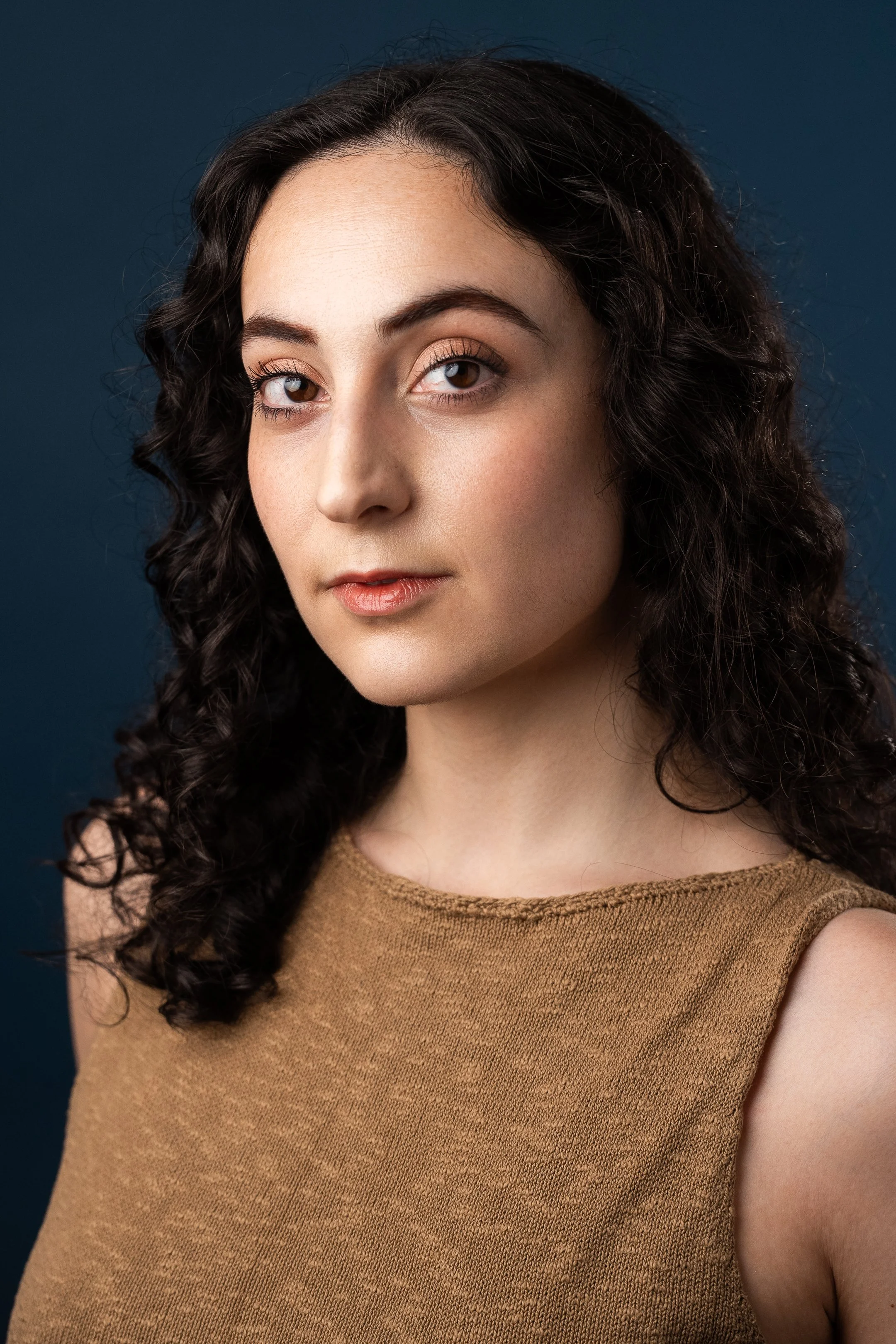 A portrait of a young woman with dark curly hair, light skin, wearing a sleeveless brown top, looking at the camera against a dark blue background.