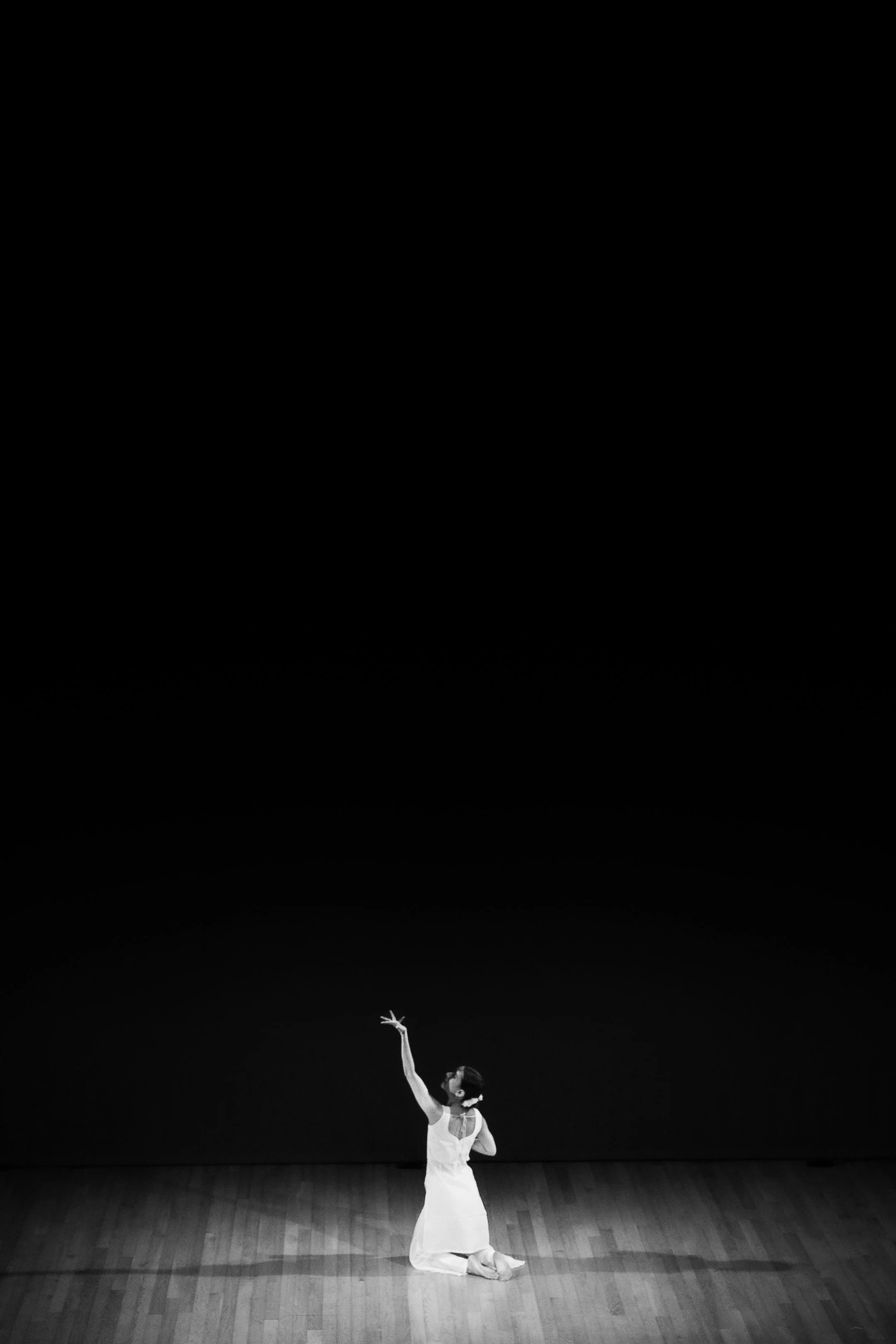 A ballerina dressed in a white dress and ballet shoes performing a dance on a wooden stage, with a dark background and ceiling.