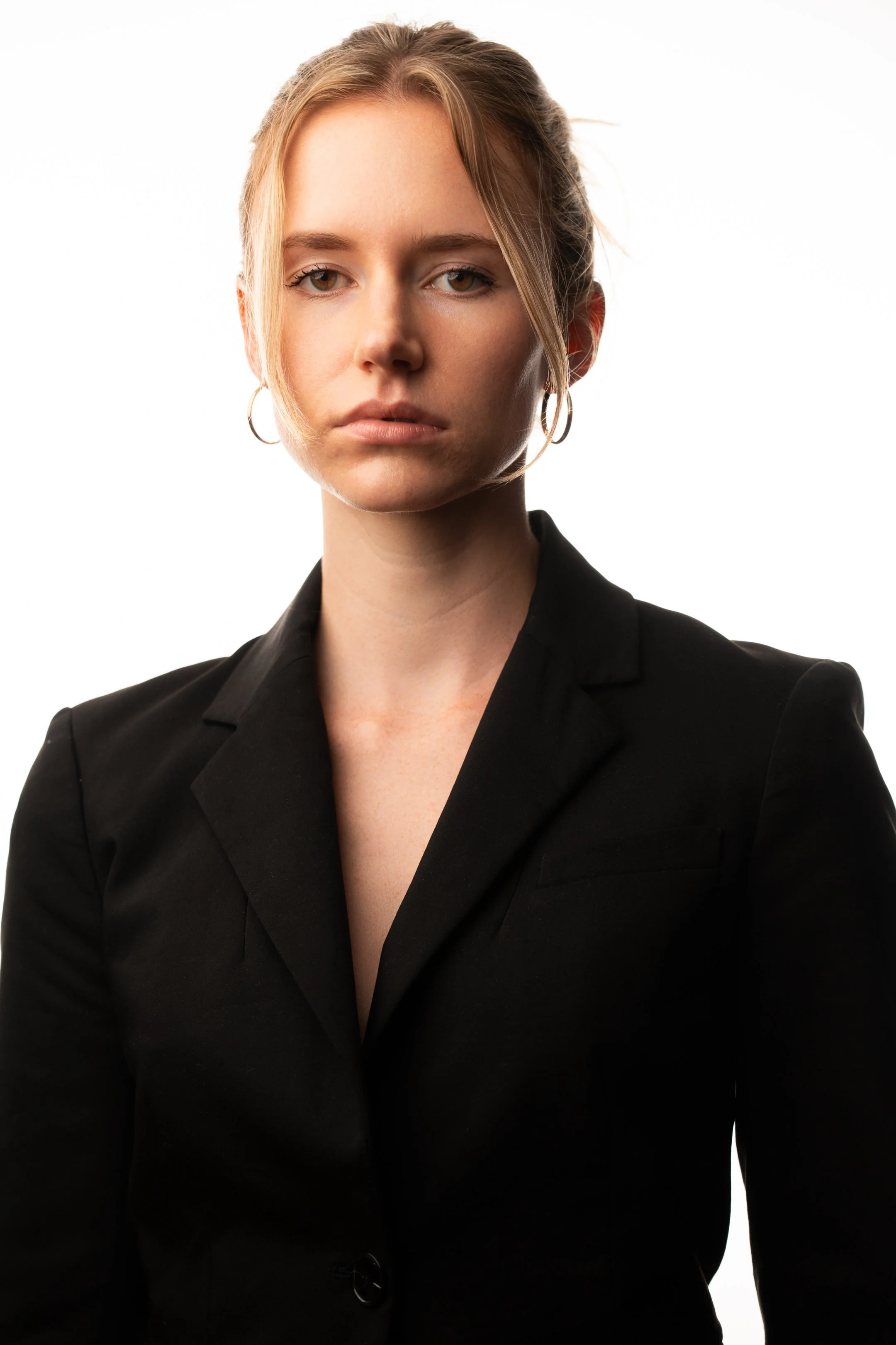 A woman with blonde hair wearing a black blazer and hoop earrings looking directly at the camera against a white background.