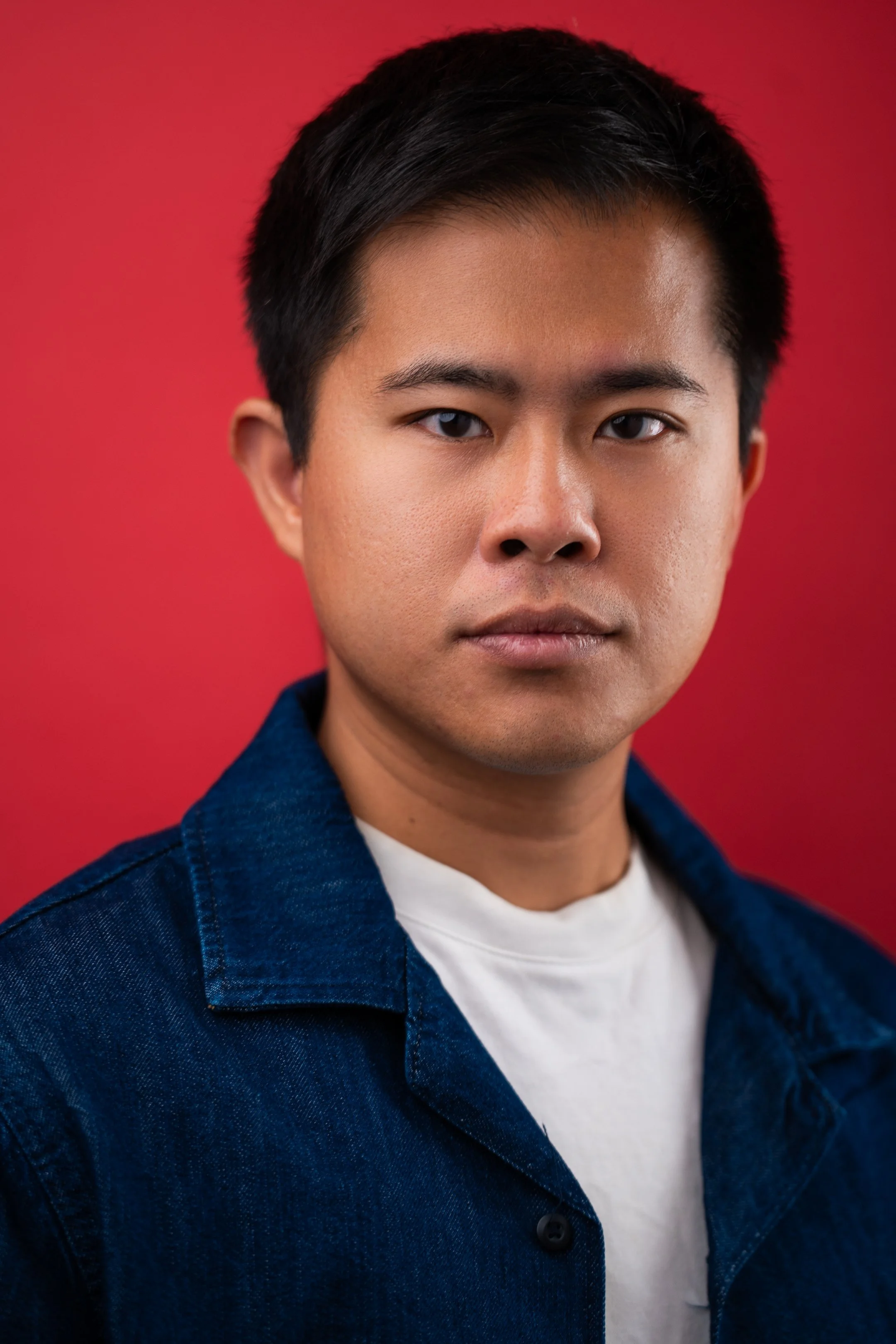 Close-up portrait of a young man with short black hair, wearing a denim jacket over a white t-shirt, standing against a red background.