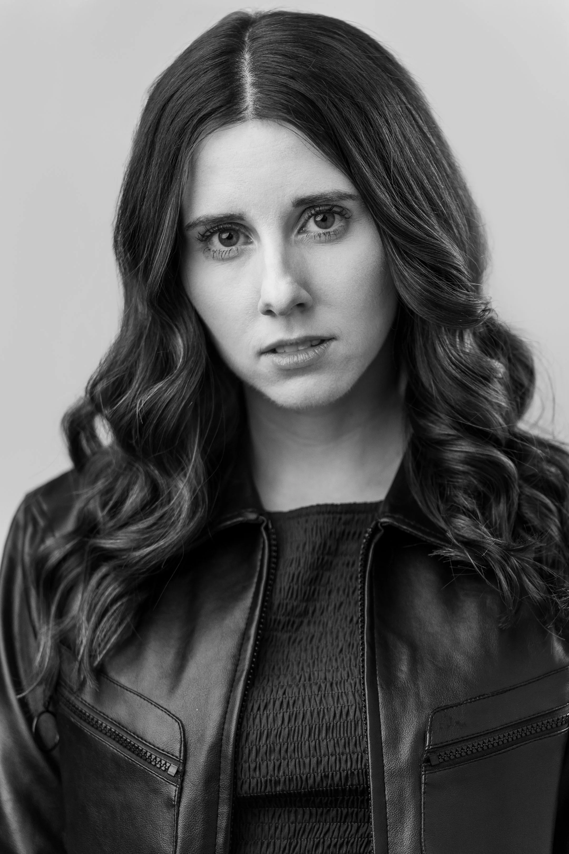Black and white portrait of a woman with wavy hair wearing a leather jacket and a textured top, looking directly at the camera.