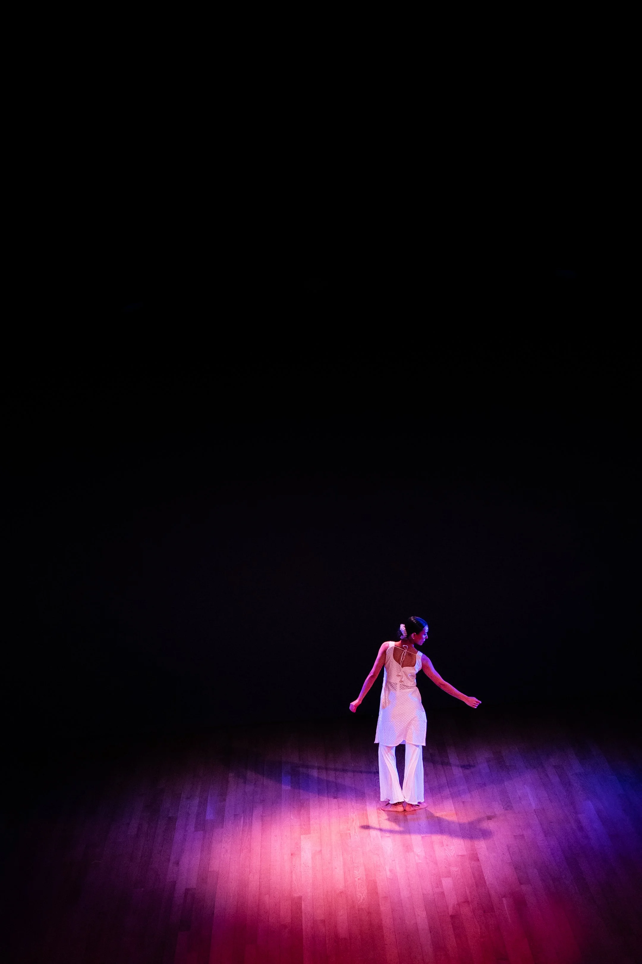 A dancer performs alone on a dimly lit stage with a wooden floor, wearing a white and pink costume, illuminated by a spotlight.