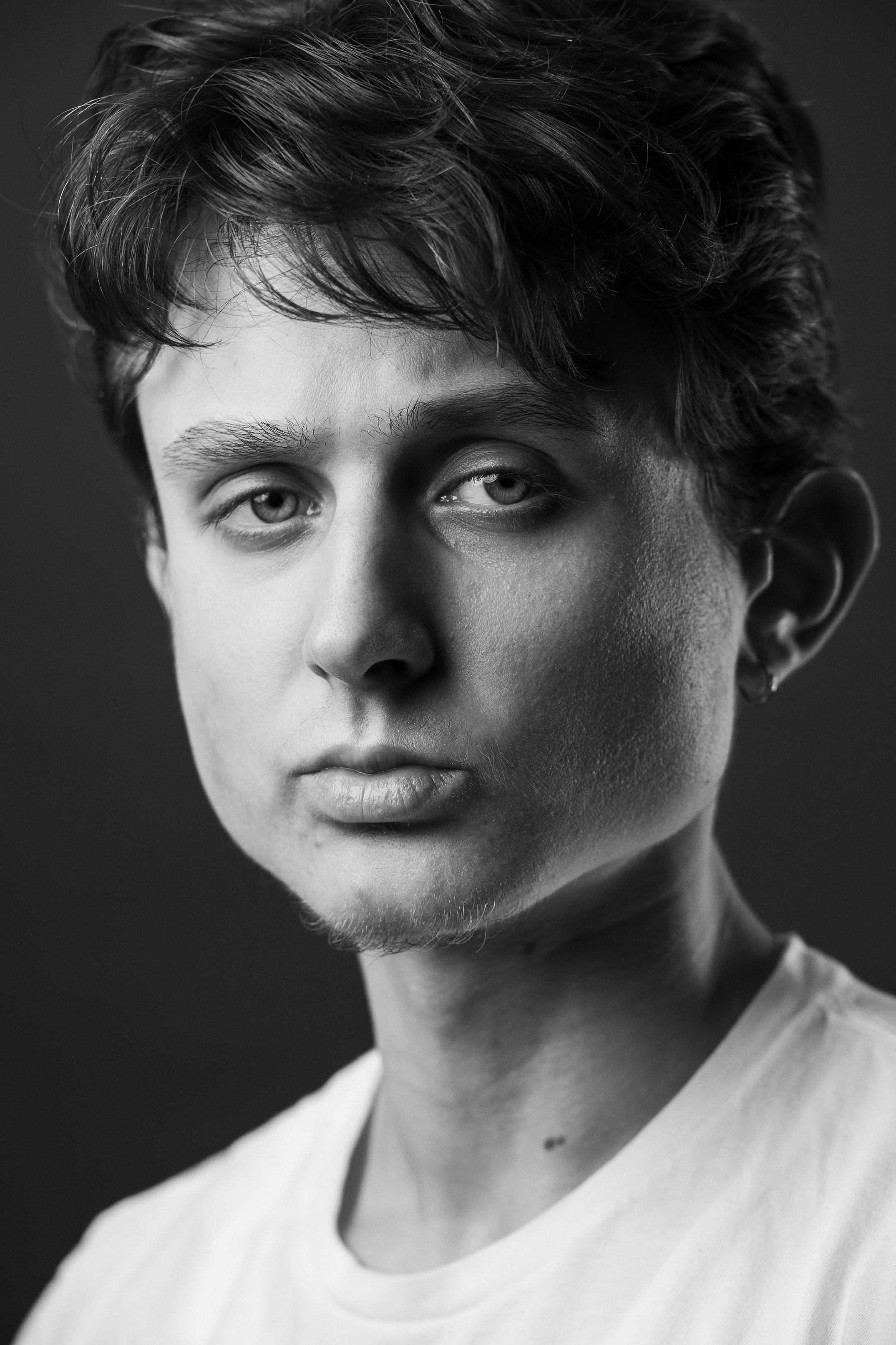 A close-up black and white portrait of a young man with short, wavy hair, light eyes, and a slight beard and mustache, wearing a plain T-shirt.
