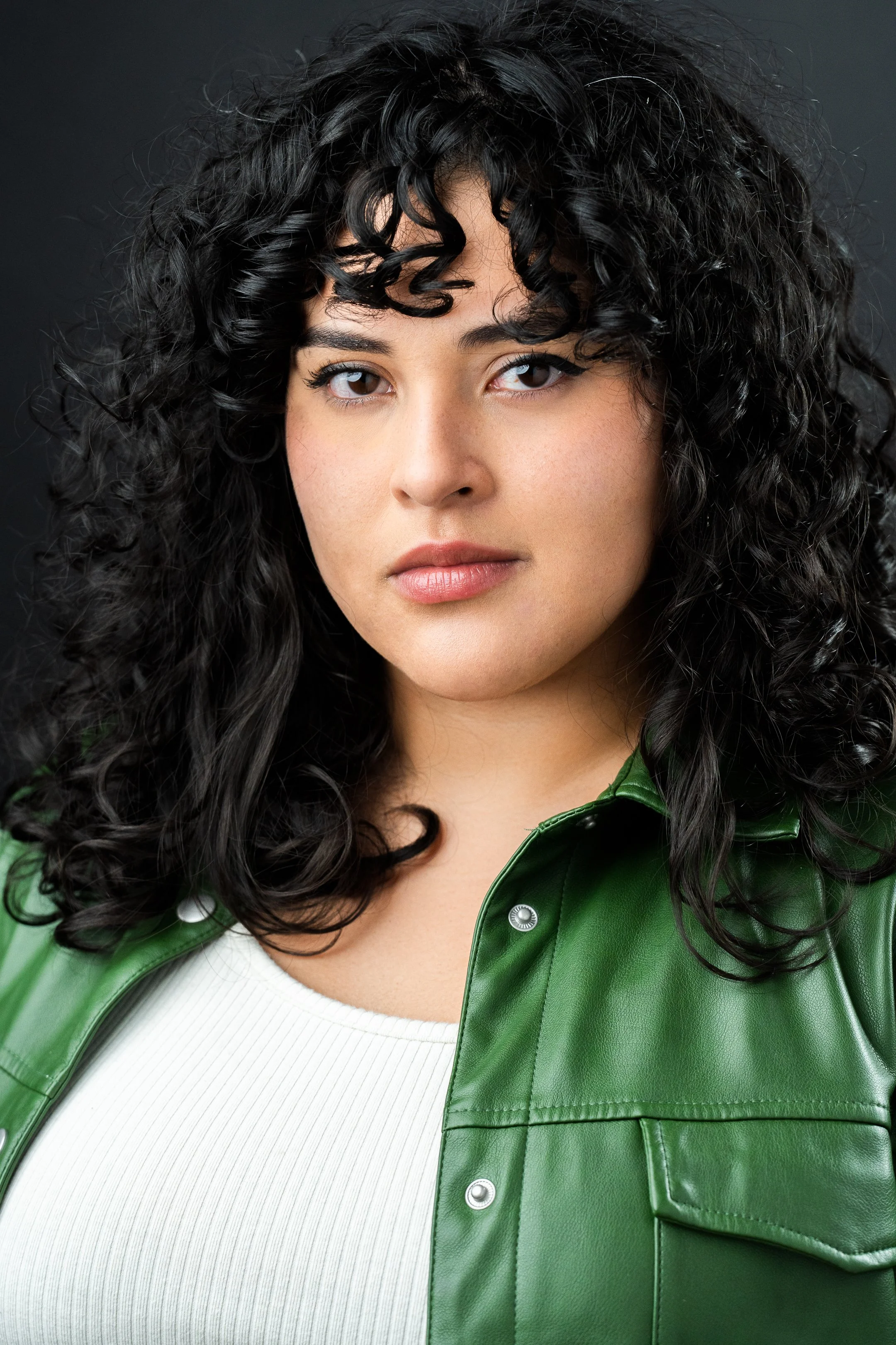 A woman with curly black hair and fair skin wearing a white top and green leather jacket, looking directly at the camera against a dark background.