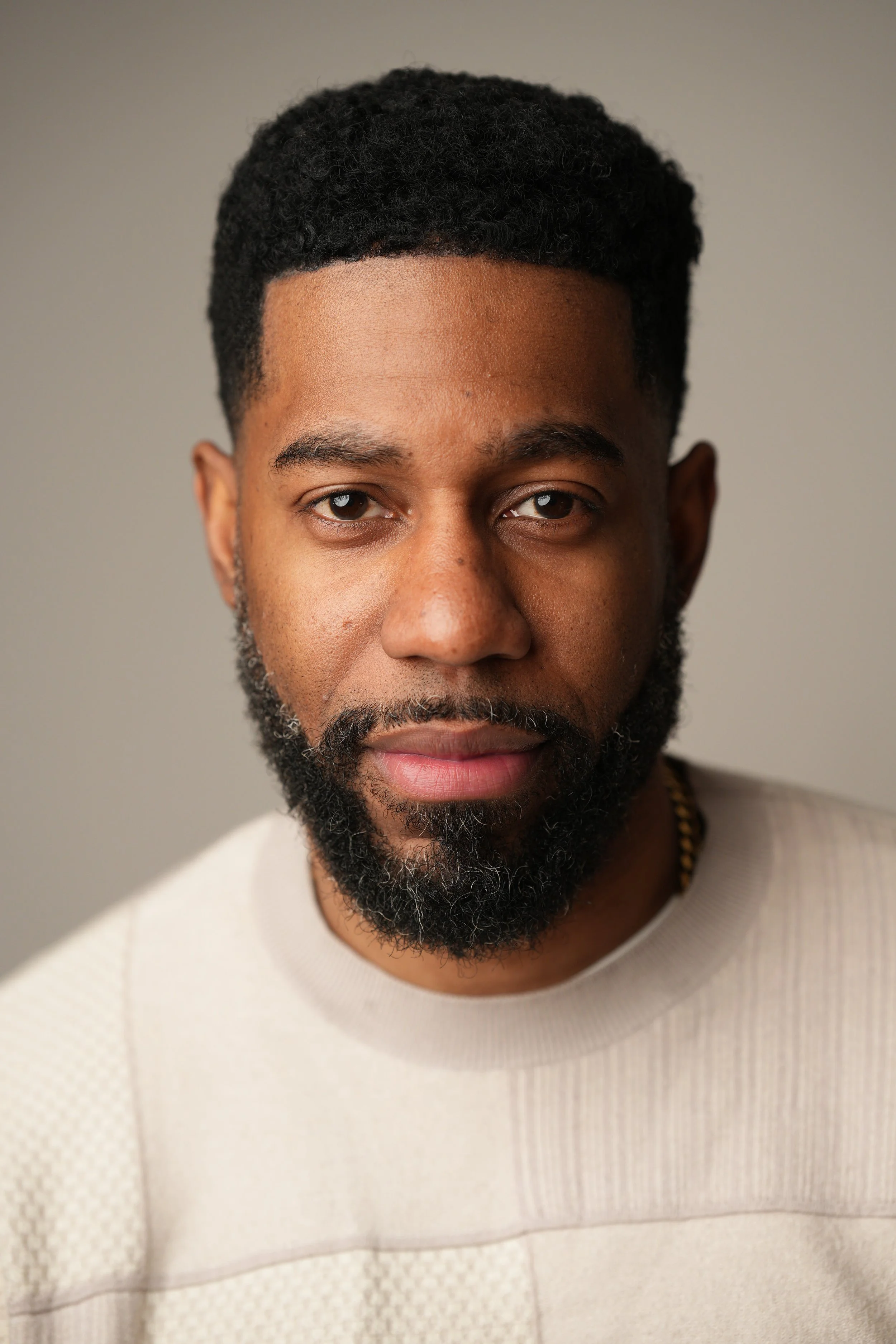 Close-up portrait of a man with a beard, short curly hair, wearing a cream-colored sweater, looking directly at the camera against a neutral background.