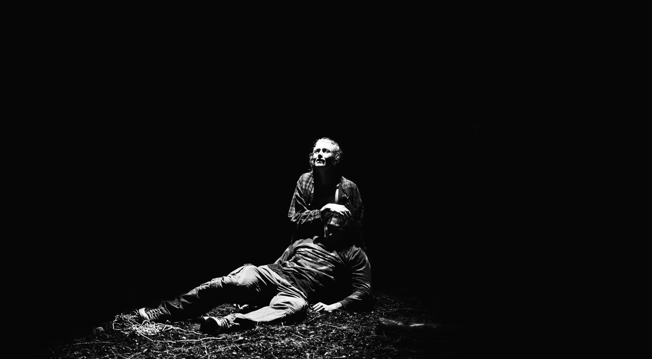 A theatrical black-and-white scene with a woman kneeling and looking upwards, illuminated by a spotlight, while a man lies on the ground with his face turned away, on a dark stage with textured ground.