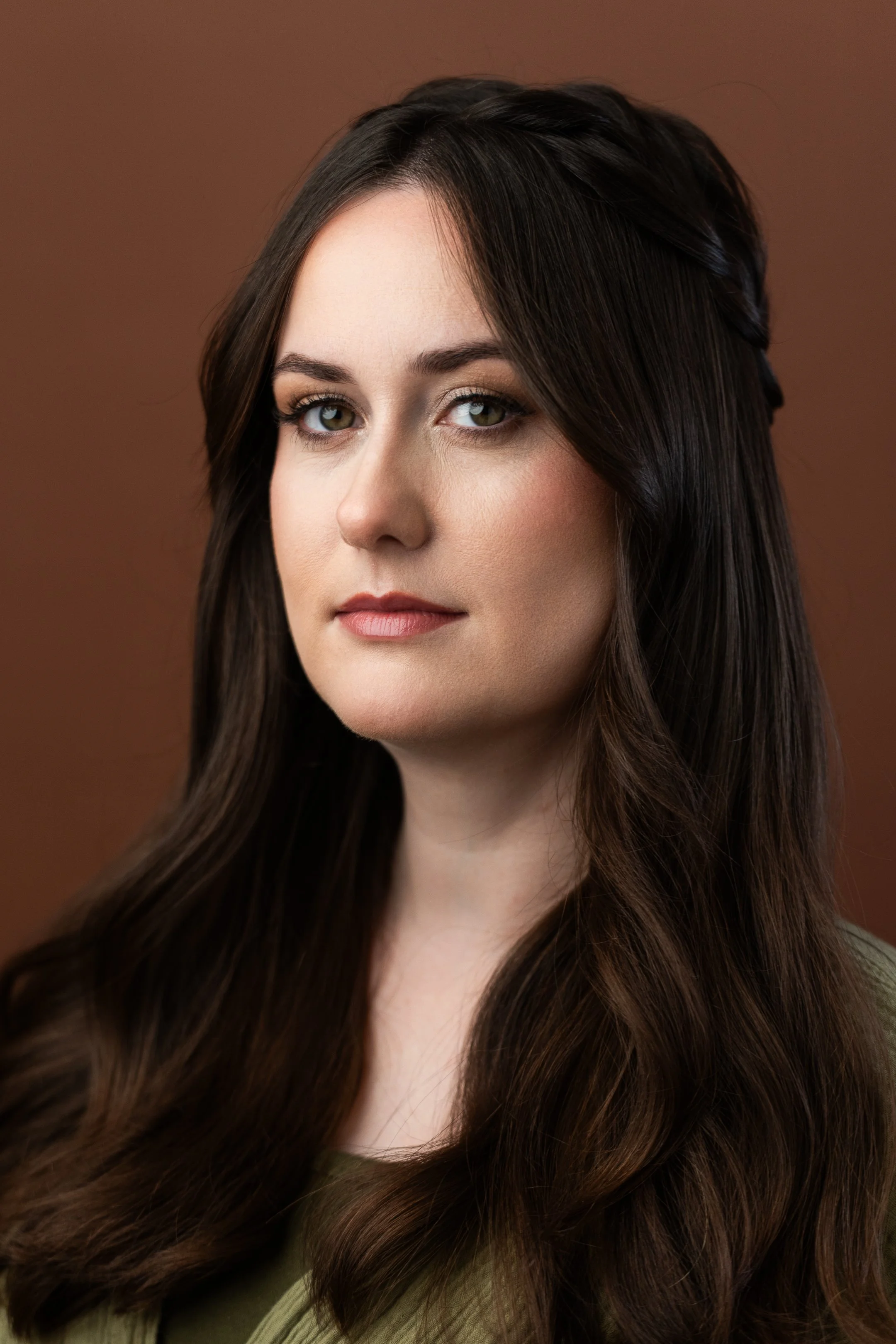 Close-up portrait of a woman with long, dark brown hair and blue eyes, wearing a green top, against a plain brown background.
