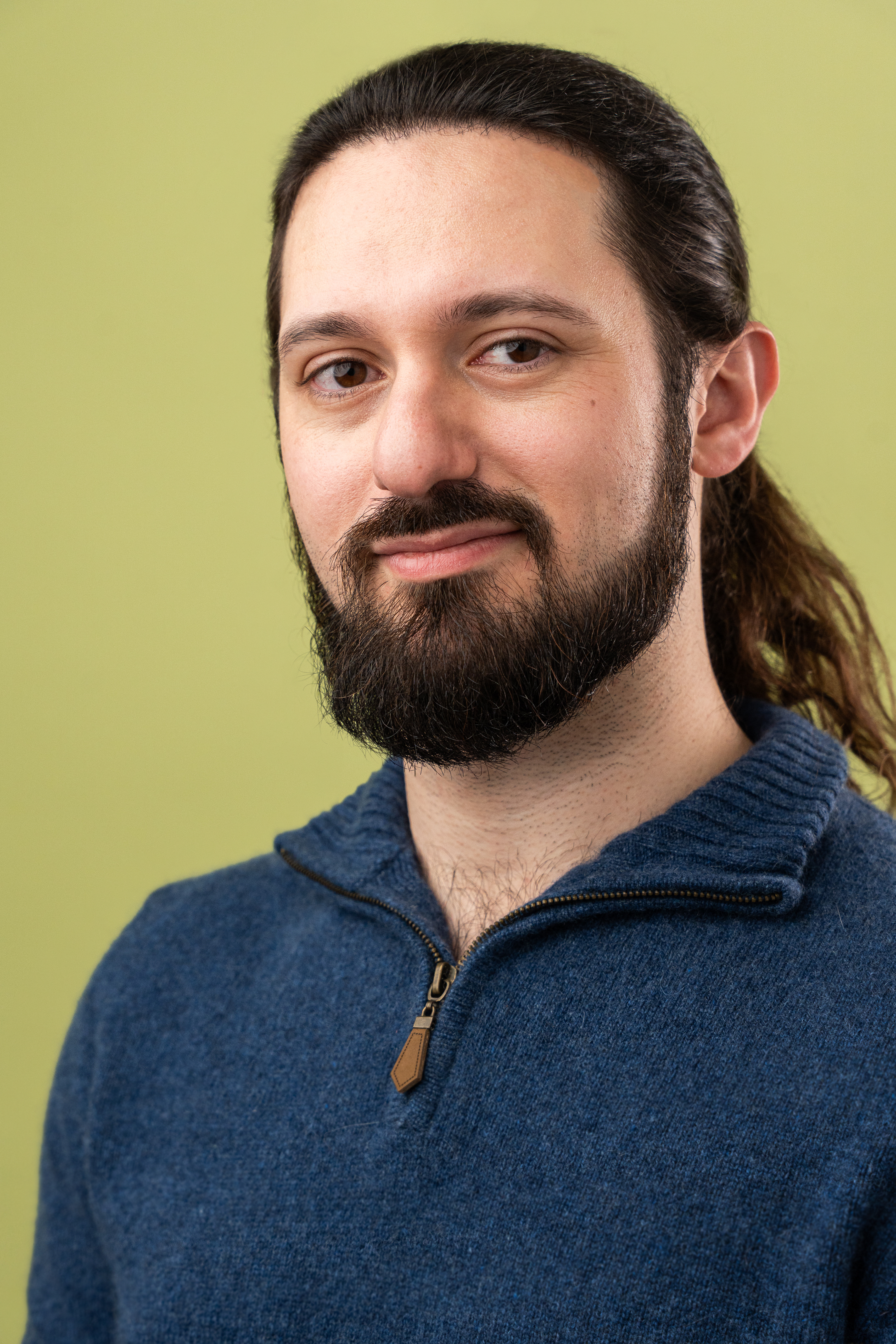Close-up portrait of a young man with long brown hair, a beard, and mustache, wearing a blue sweater with a zipper, against a light green background.