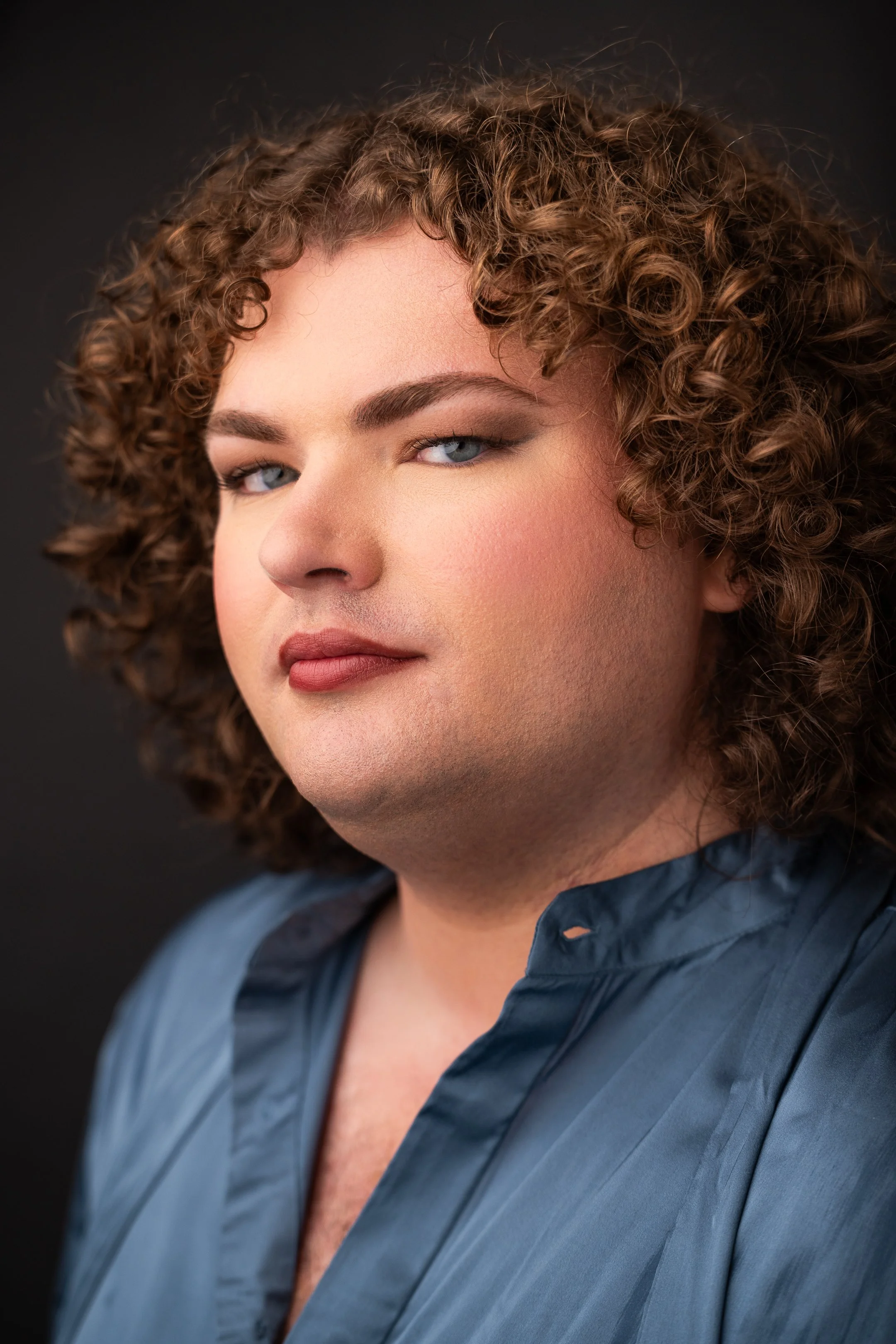 A close-up portrait of a person with curly brown hair, light makeup, and blue eyes, wearing a dark blue satin shirt, against a dark background.