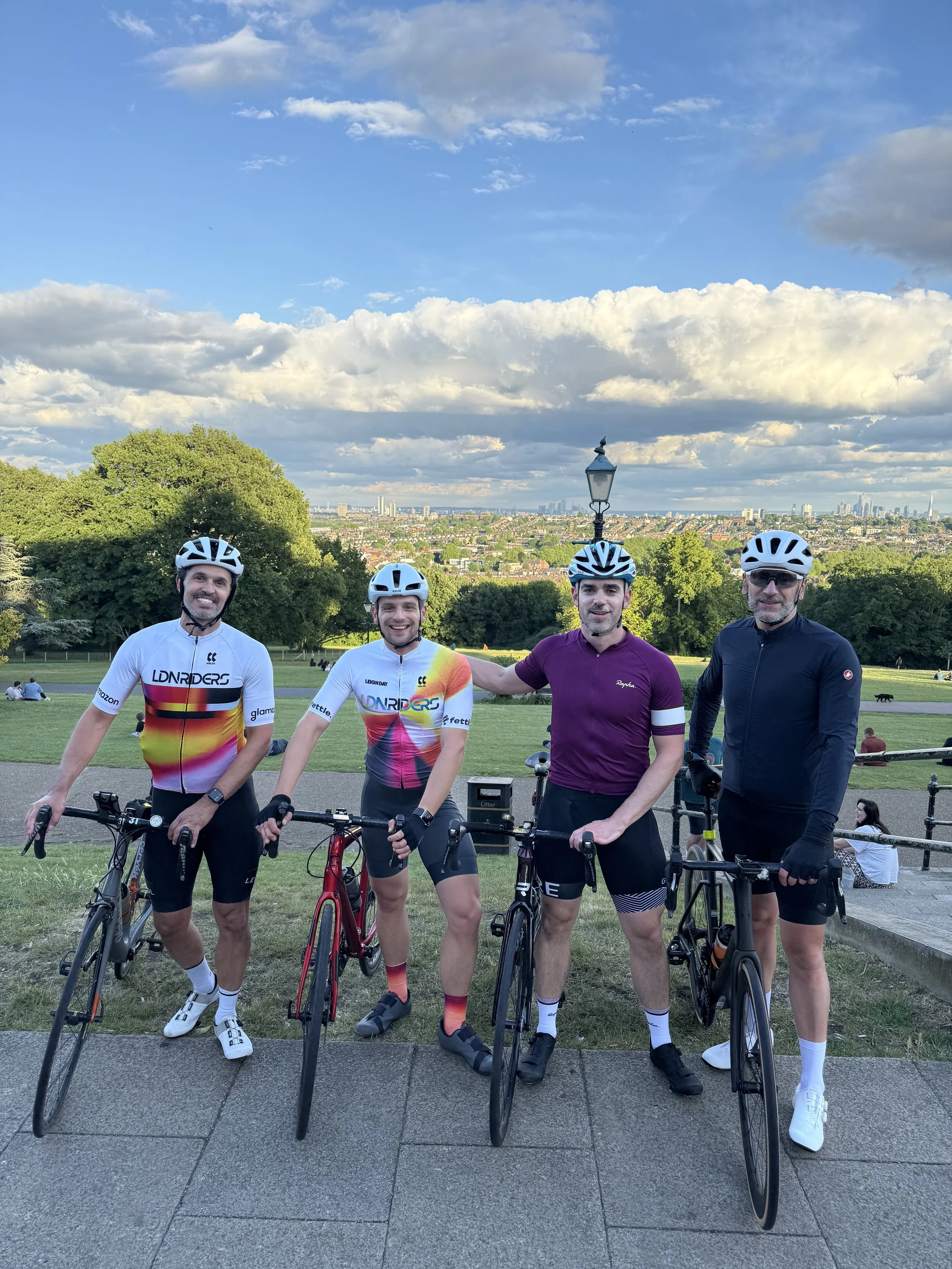Four men in cycling gear standing with their bikes in a park with trees, a lamppost, and a city skyline in the background.