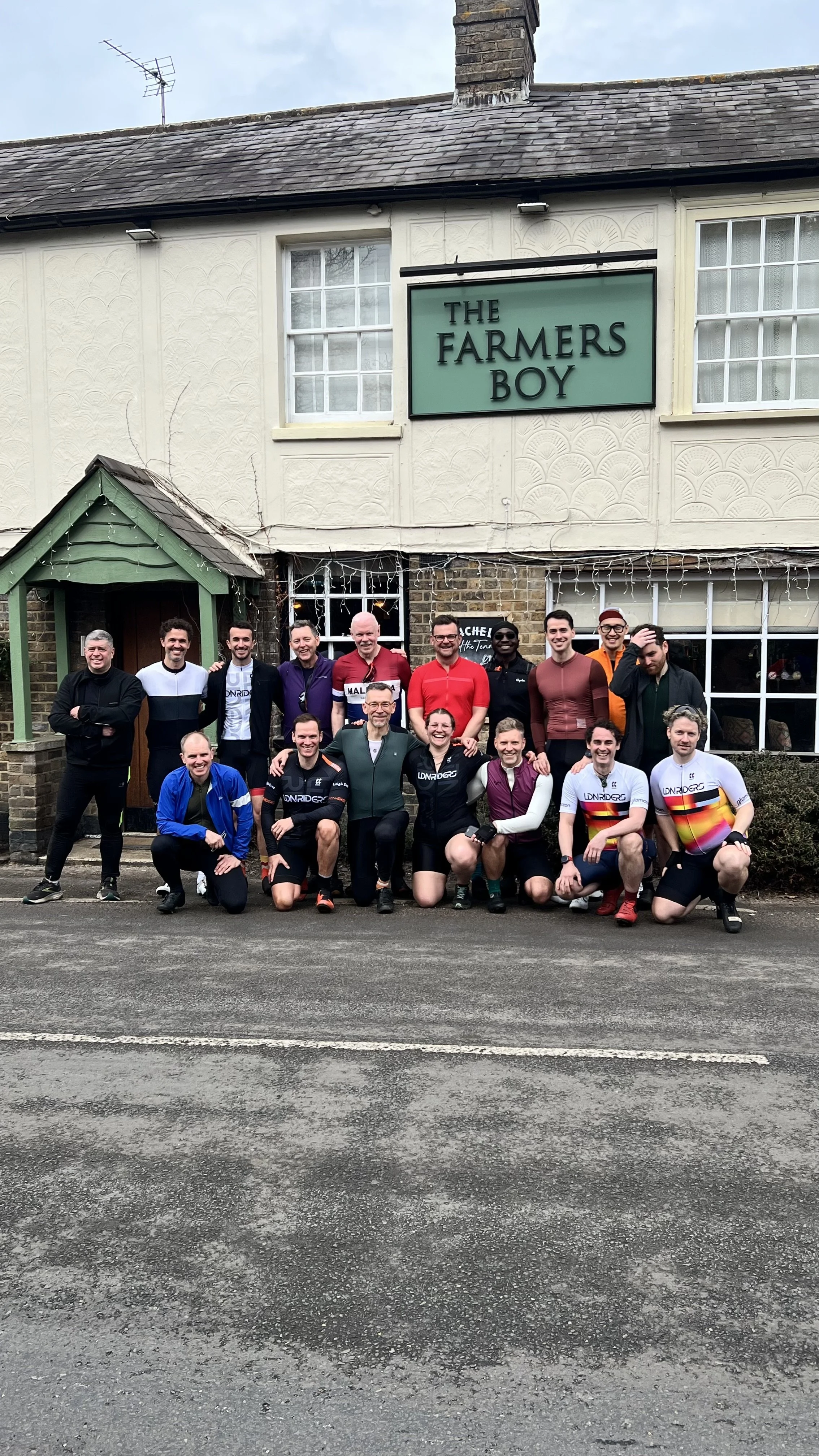 A group of people posing outside a pub called The Farmers Boy, with some dressed in cycling gear.