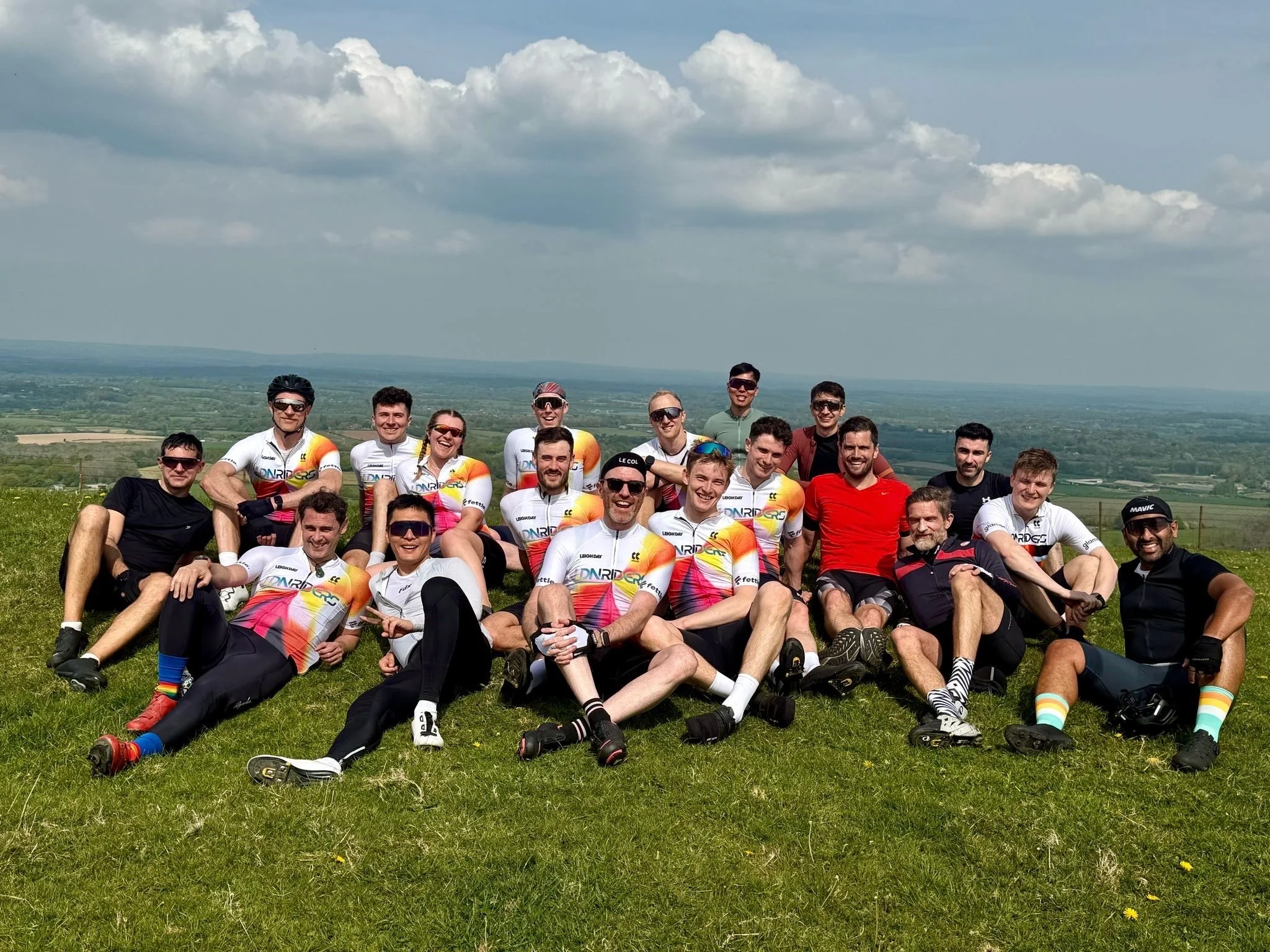 Group of cyclists in sports gear sitting on grass outdoors with a scenic landscape of open fields and hills under a partly cloudy sky.