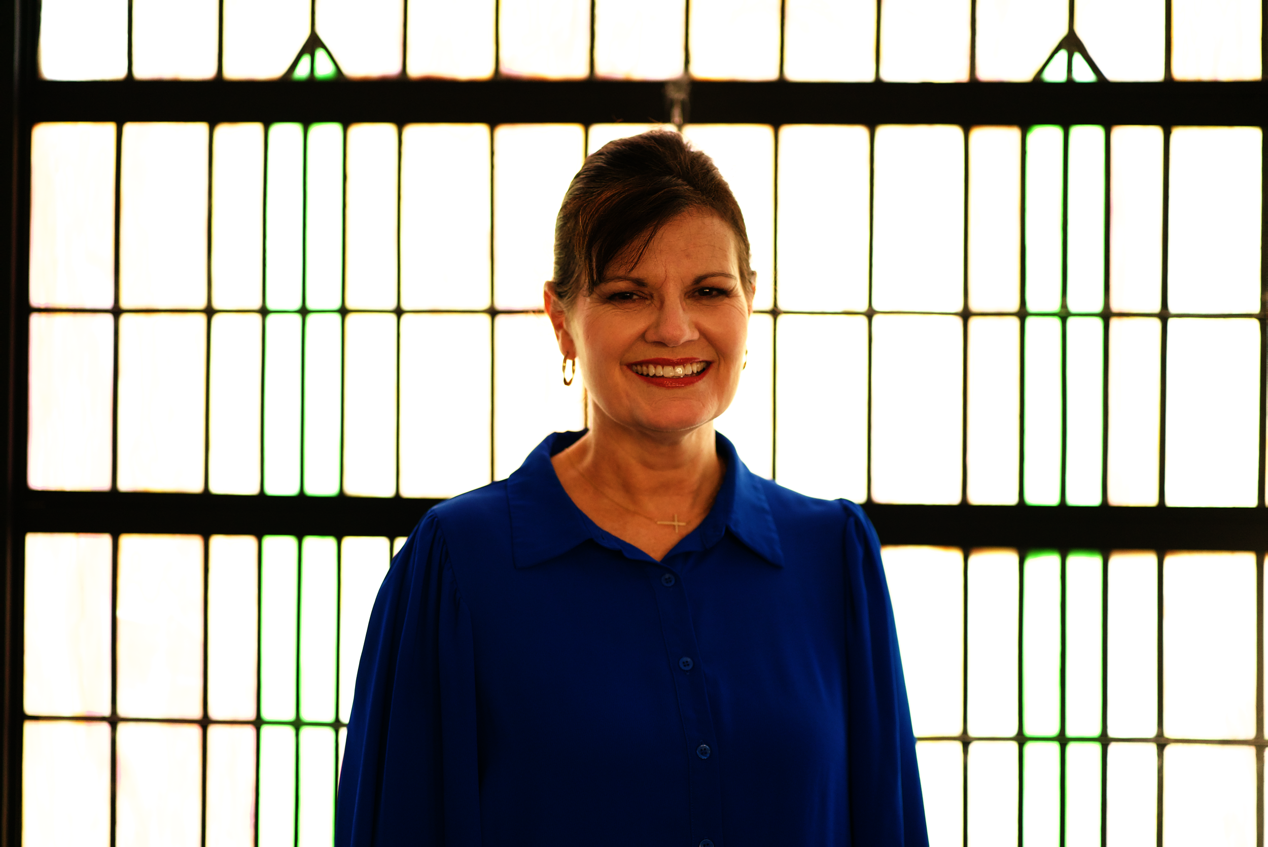 Woman wearing glasses and a blue embroidered top, standing outside in a garden or park in front of a brick building.