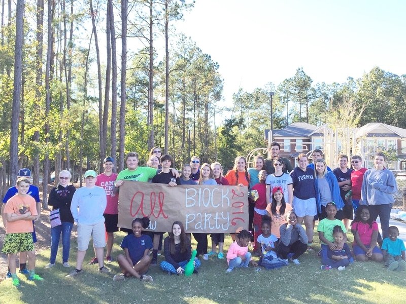 A large group of people of various ages standing outdoors in a park-like setting. They are holding a sign that says "All Block Party 2016." The group includes adults and children, and several of them are wearing casual clothing. There are trees, a playground, and residential buildings in the background, indicating a community gathering.