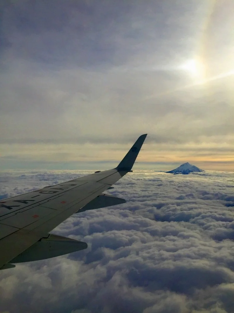 Airplane wing flying above clouds with a mountain peak in the distance and a cloudy sky with sun.