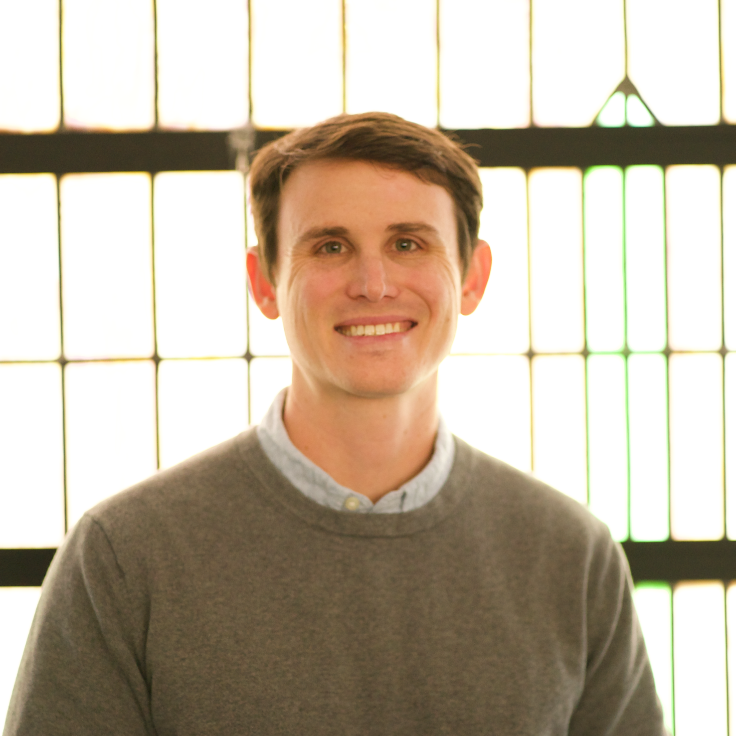 A person in a white shirt standing outside with a blurred background of greenery and a red brick building.