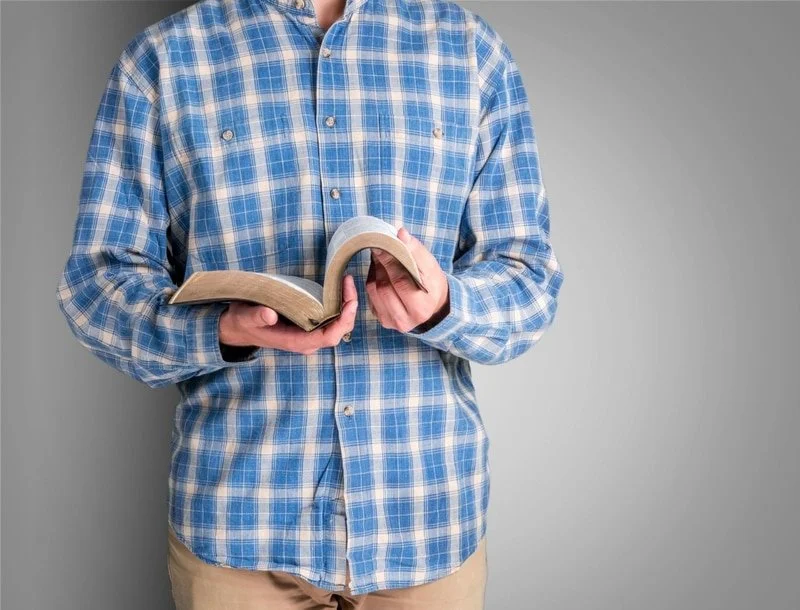 Person in blue plaid shirt holding an open book against a gray background.