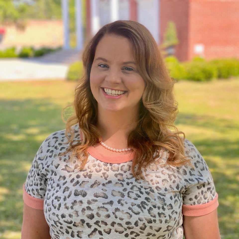 Smiling woman with wavy hair wearing a leopard print top and a pearl necklace, standing in a sunny outdoor setting with trees and a building in the background.