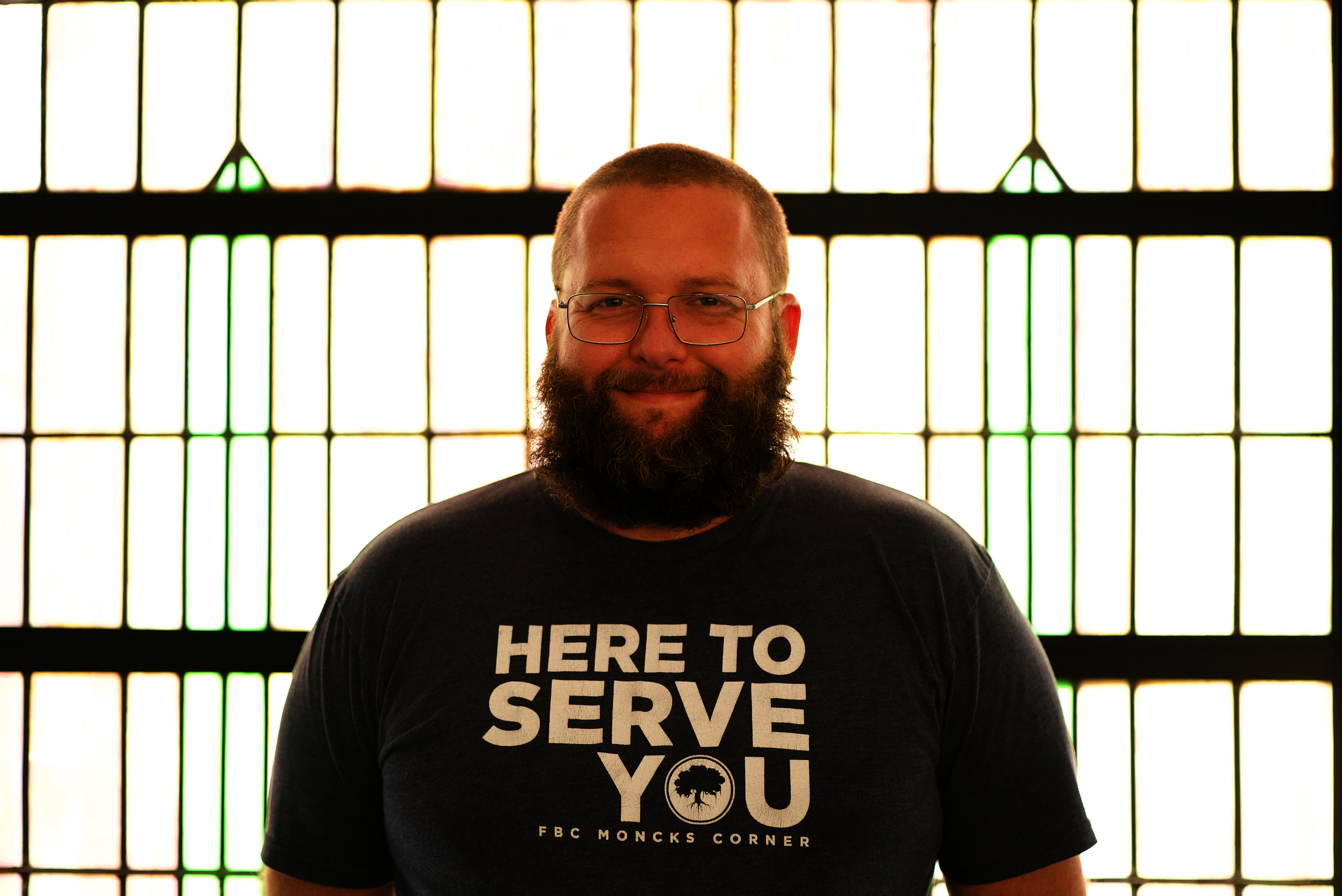 Man with glasses, beard, and plaid shirt standing outdoors in front of a building with columns.