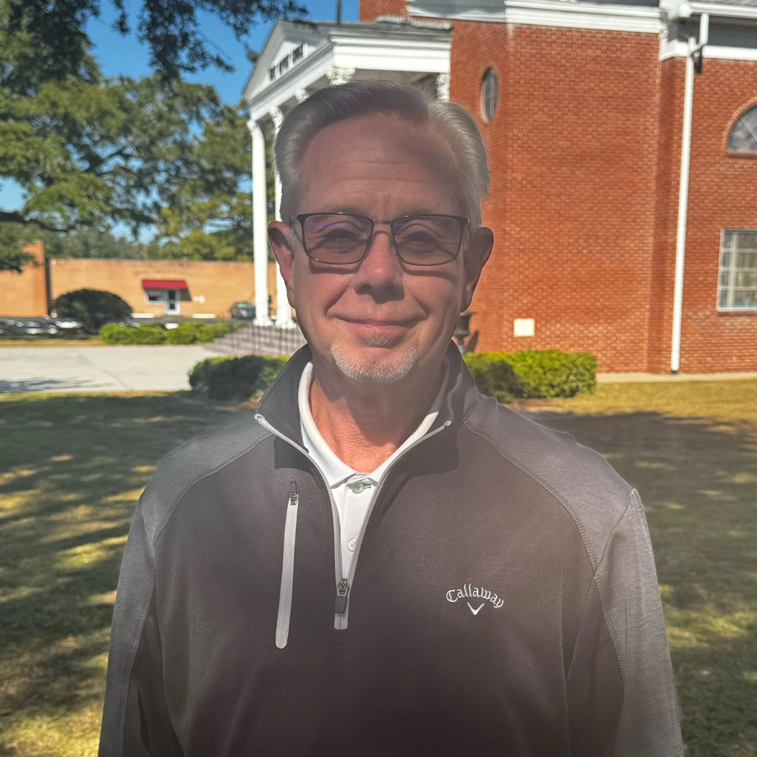 Man wearing glasses and a Callaway jacket standing in front of a red brick building with white columns.