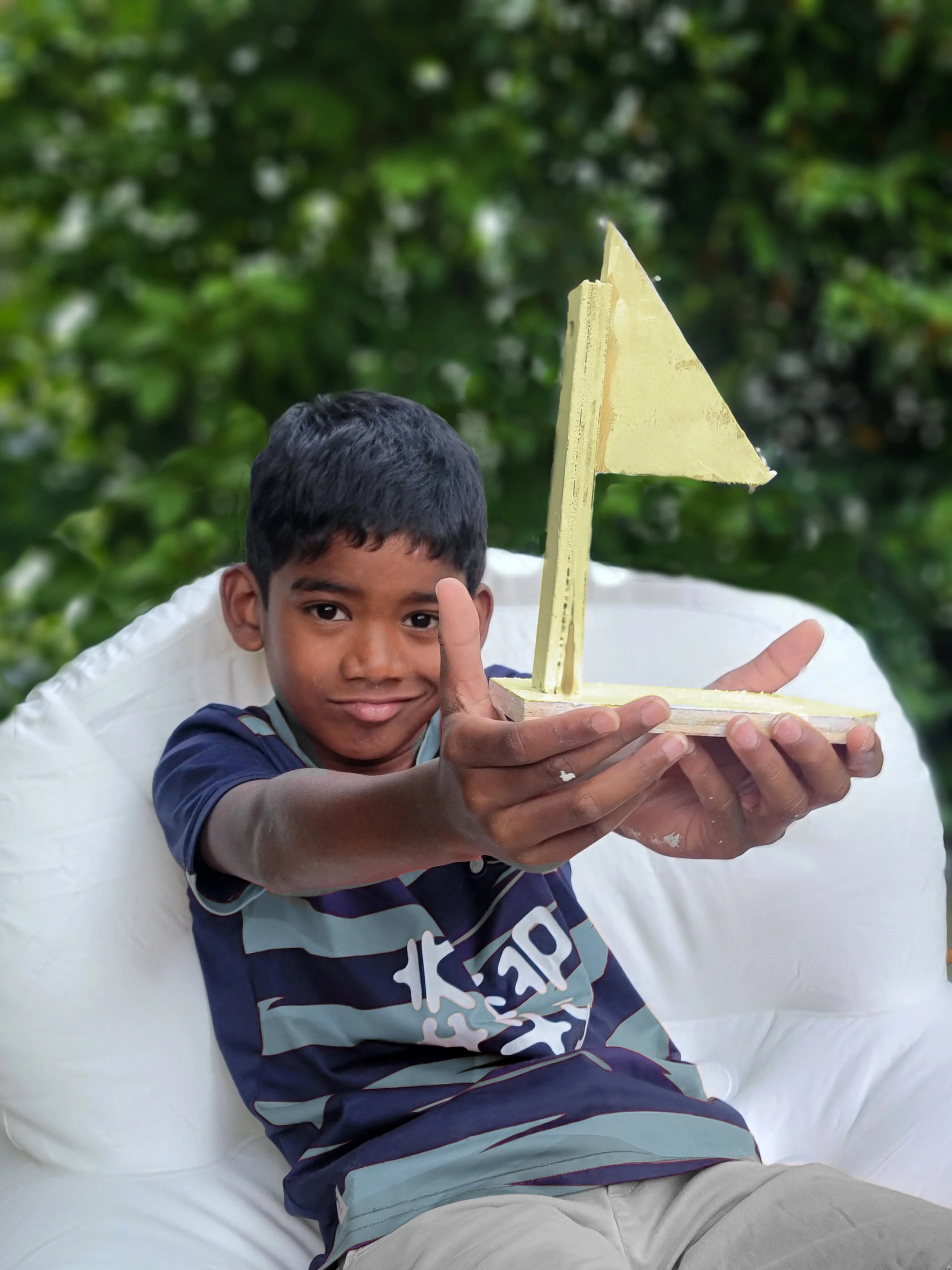 Un garçon de 8 ans tenant une maquette de bateau en bois peint en jaune, positionné devant un fond de feuillages verts.