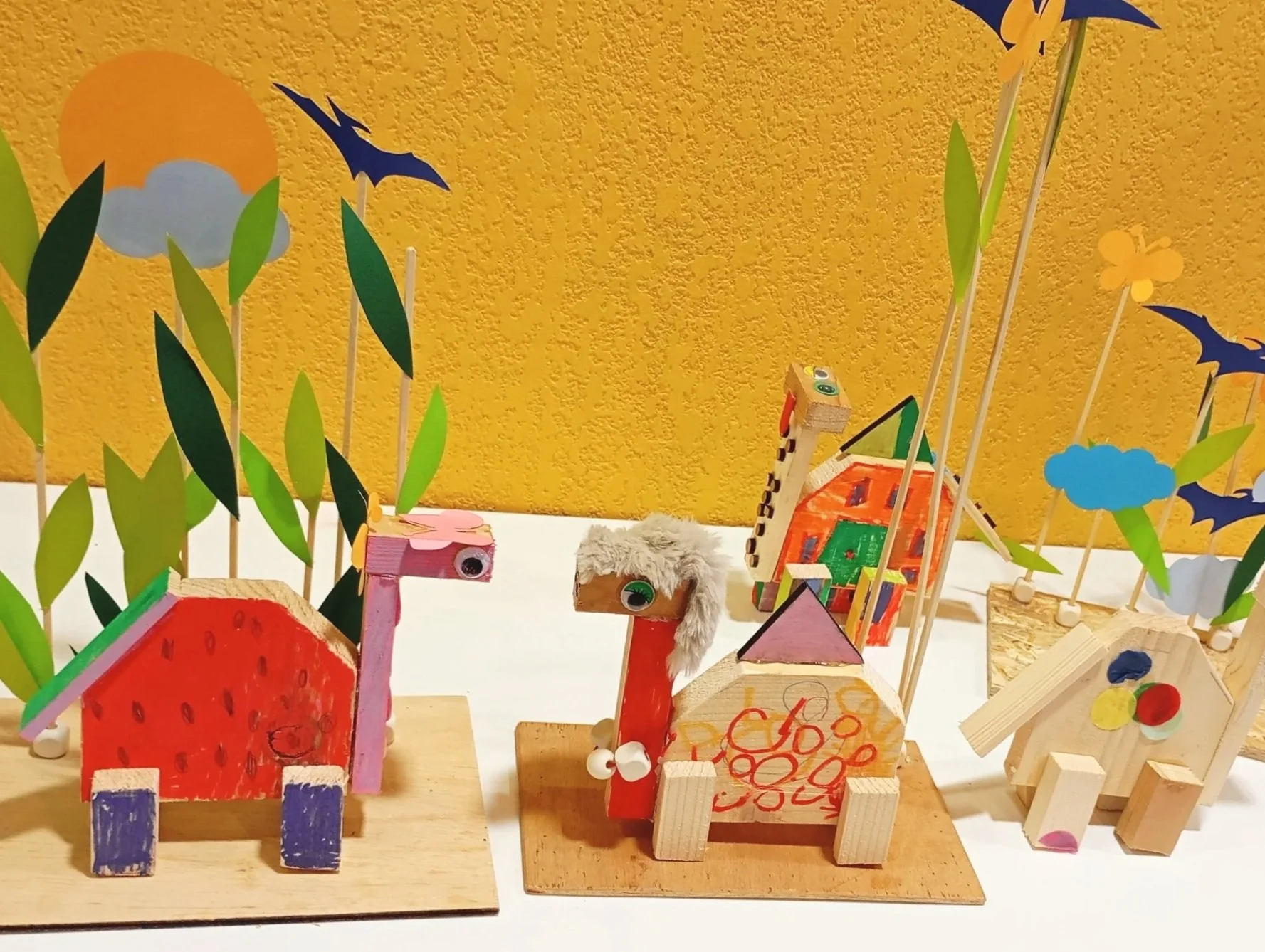 Petits animaux ou dinosaures en bois décorés avec de la laine et des matériaux de récupération, entourés de décorations en papier représentant un soleil, des nuages, des feuilles, et des chauves-souris, avec un fond jaune.