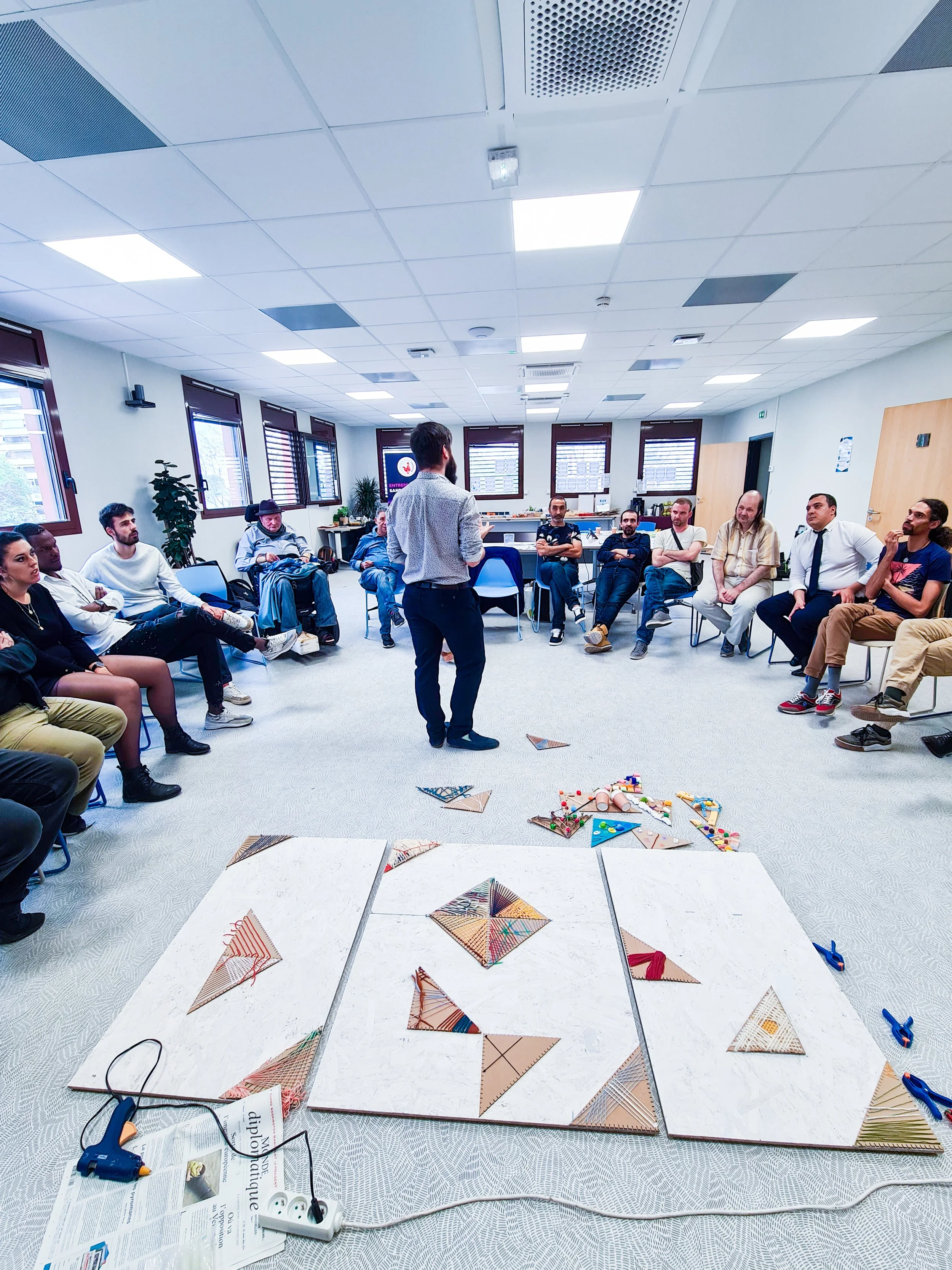 Une personne donne une présentation à un groupe de personnes assises en demi-cercle dans une salle, avec des papiers, des ciseaux et des objets colorés sur le sol. Atelier de Team Building en entreprise.