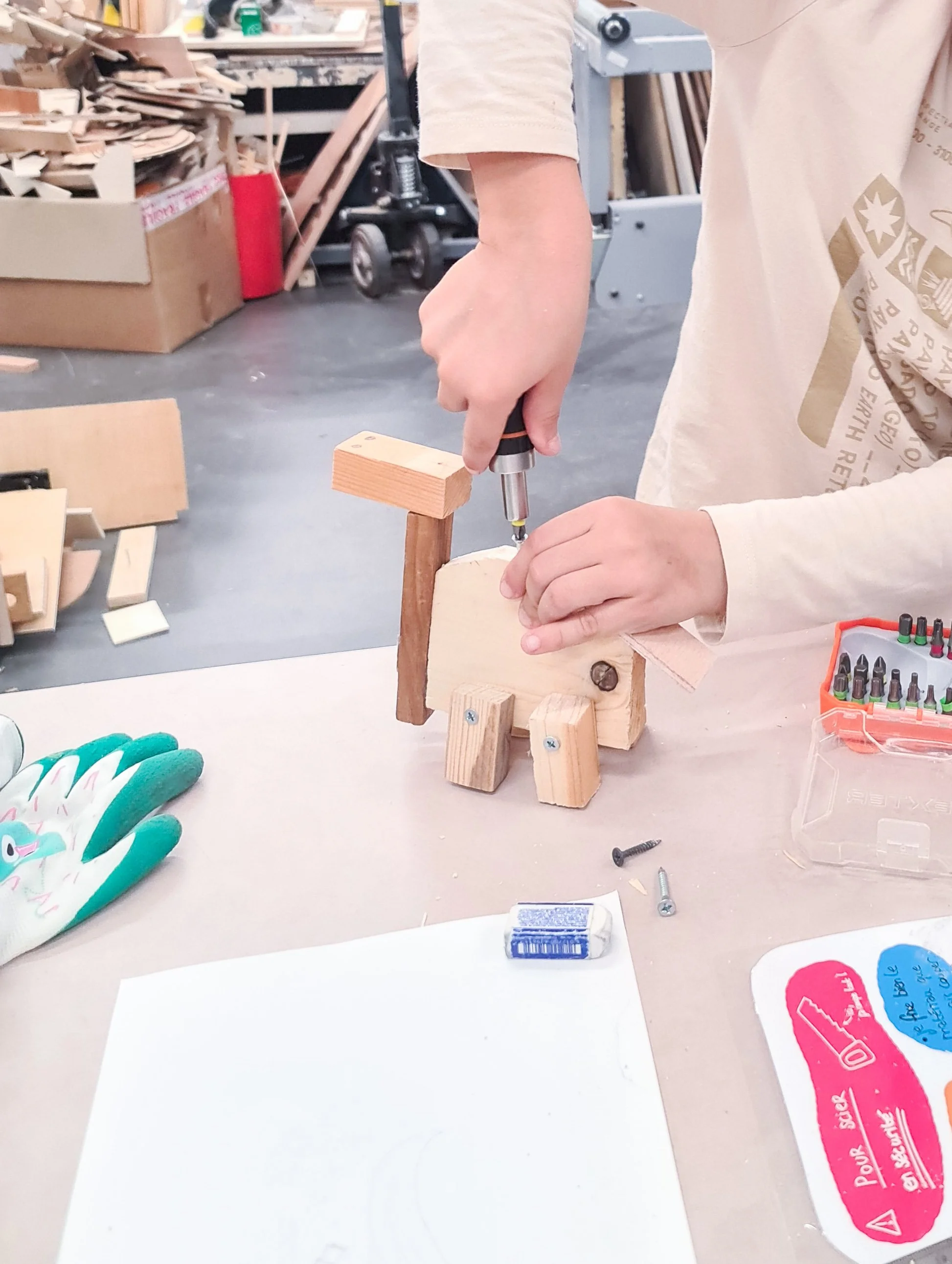 Un enfant assemble une sculpture en bois en utilisant un tournevis, dans un atelier de menuiserie ou de bricolage, avec divers outils et matériaux autour.