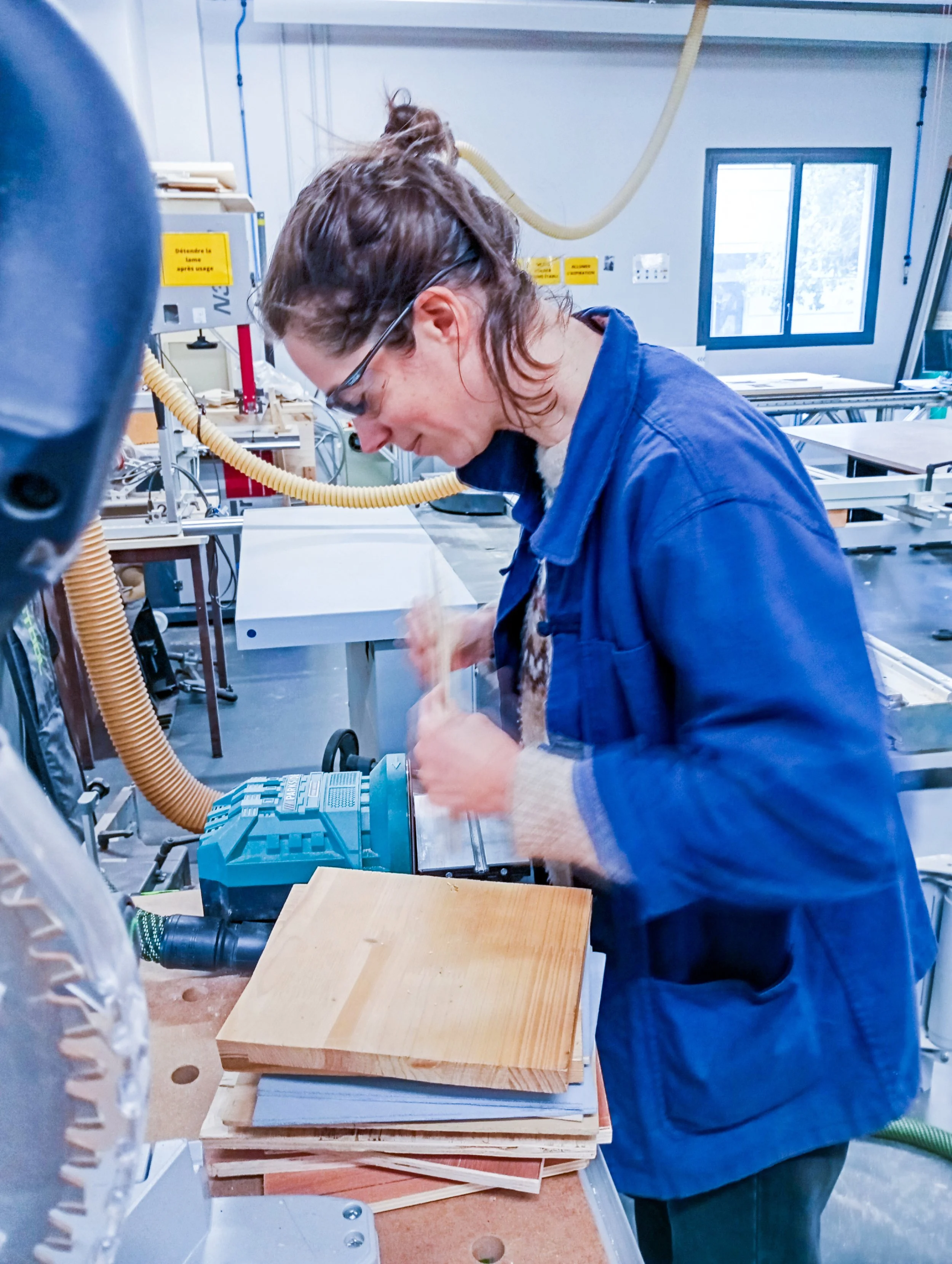 Une femme travaille dans un atelier de menuiserie, utilisant une machine à bois pour couper ou façonner une pièce de bois. Elle porte des lunettes de protection et un manteau bleu.